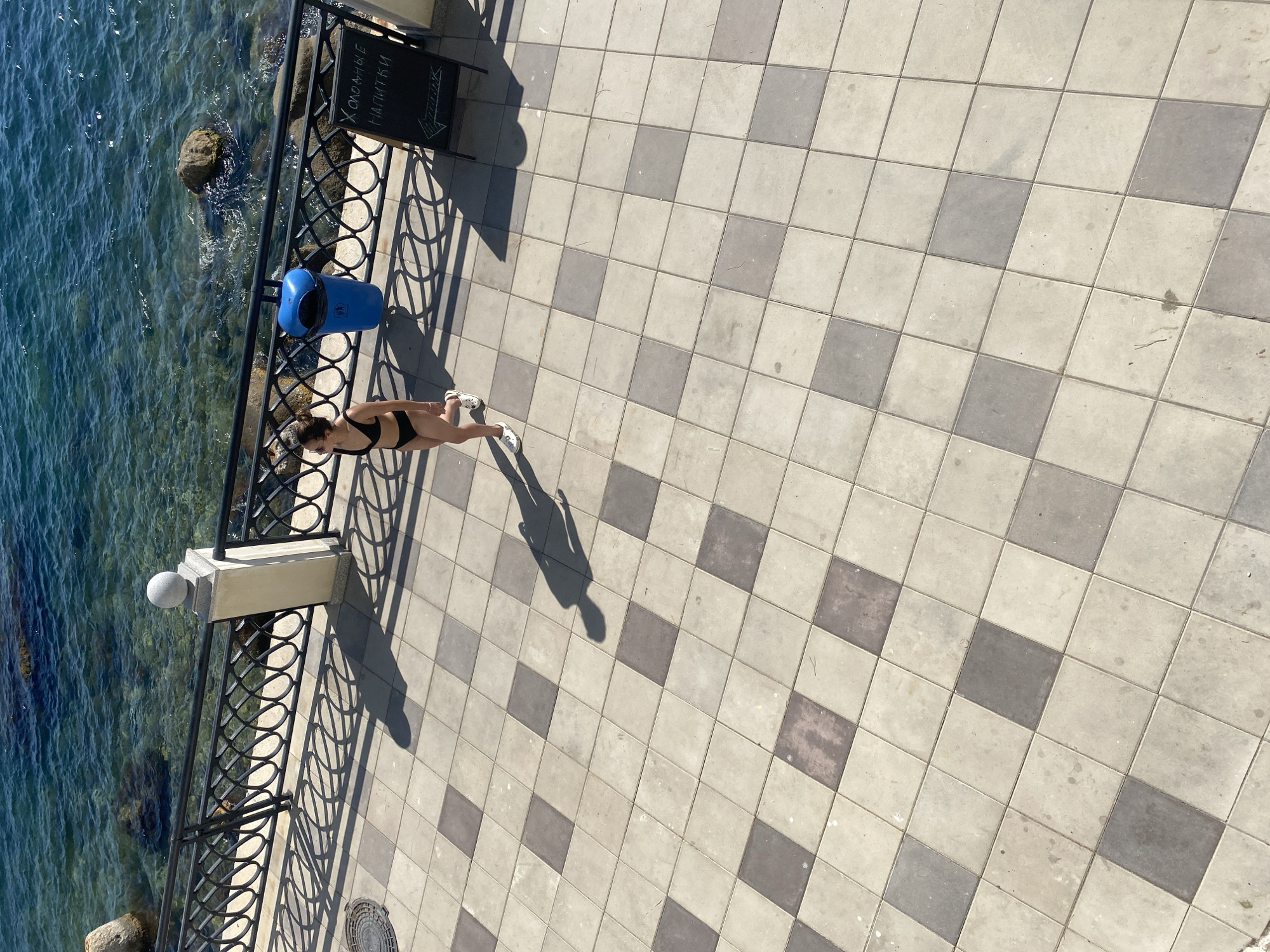 A young woman in a black bikini walks across a checkered tiled promenade next to the blue sea. She has dark hair pulled back and is wearing white sneakers. A black metal railing separates the promenade from the water. A blue trash can is visible near the railing. To the right, a blackboard sign displays text in Cyrillic. The sun casts shadows of the railing and the woman onto the tiles. The sea is clear and calm, with rocks visible beneath the surface.
