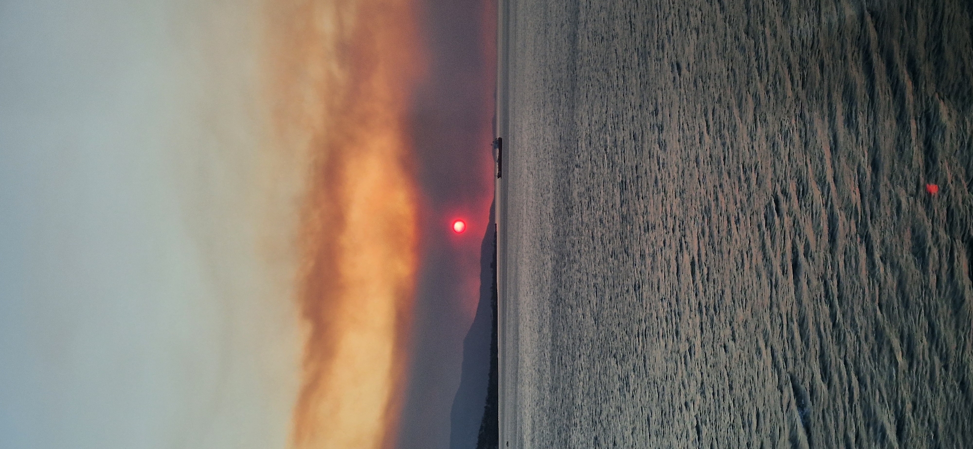 The image depicts a sunset over the sea, with a visible layer of smoke in the sky, suggesting a possible wildfire or industrial pollution. The sun, a vibrant red sphere, is partially obscured by the smoke, casting a warm glow on the clouds. In the distance, the silhouettes of mountains can be seen along the coastline. A cargo ship is positioned on the water, contributing to the maritime atmosphere. The sea itself is choppy, with visible waves reflecting the dusky light. The scene is set within the Municipal Unit of Agioi Theodoroi, Greece, a coastal area known for its industrial activity and proximity to natural landscapes.