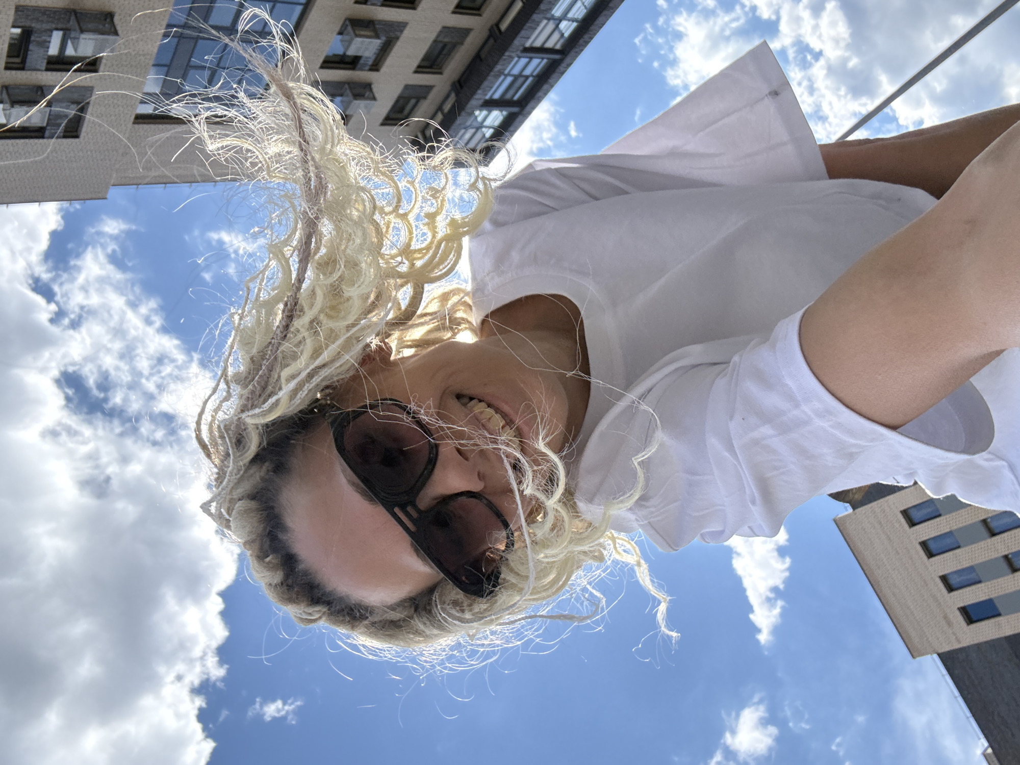 A woman wearing sunglasses and a white t-shirt looks down at the camera from an extreme low angle. Her long blonde hair is styled with braids and loose strands. A building and a blue sky with scattered clouds are visible in the background. The scene appears to be outdoors in a city.