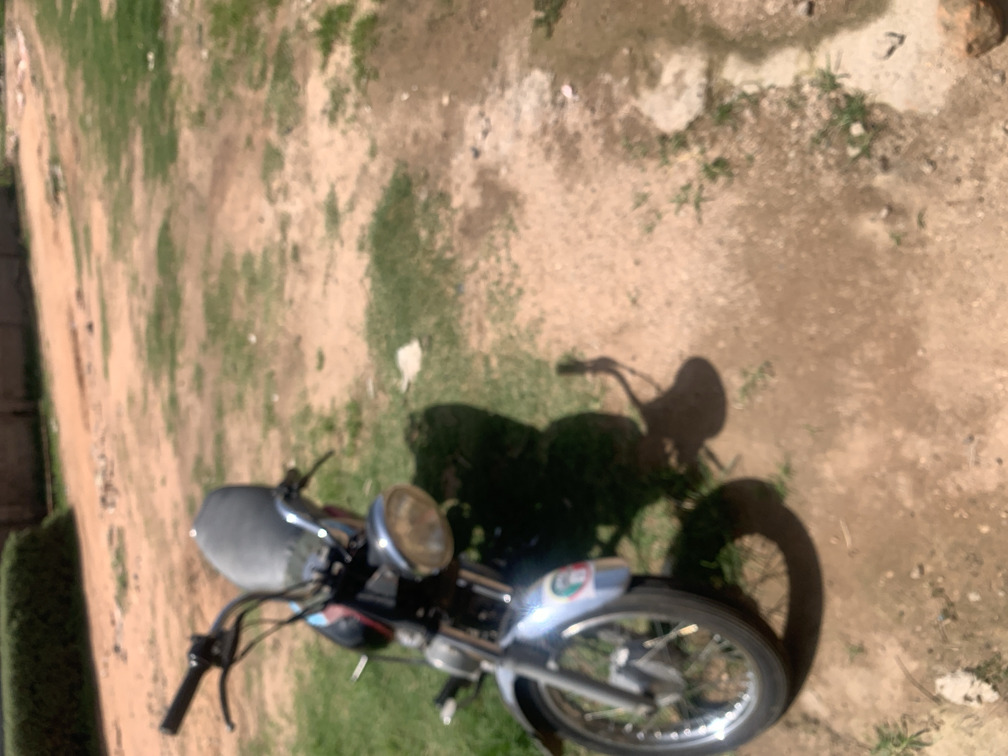 A motorcycle is parked on a dirt and grass area in Jauro Yinu, Nigeria. The motorcycle is silver and black with a large, round headlight. It is angled slightly towards the left of the frame. The ground is a mix of reddish-brown dirt and patches of green grass. In the background, there is a blurred green hedge and what appears to be a building. The scene is lit by sunlight, casting a distinct shadow of the motorcycle on the ground to its right. There are no people visible in the image.