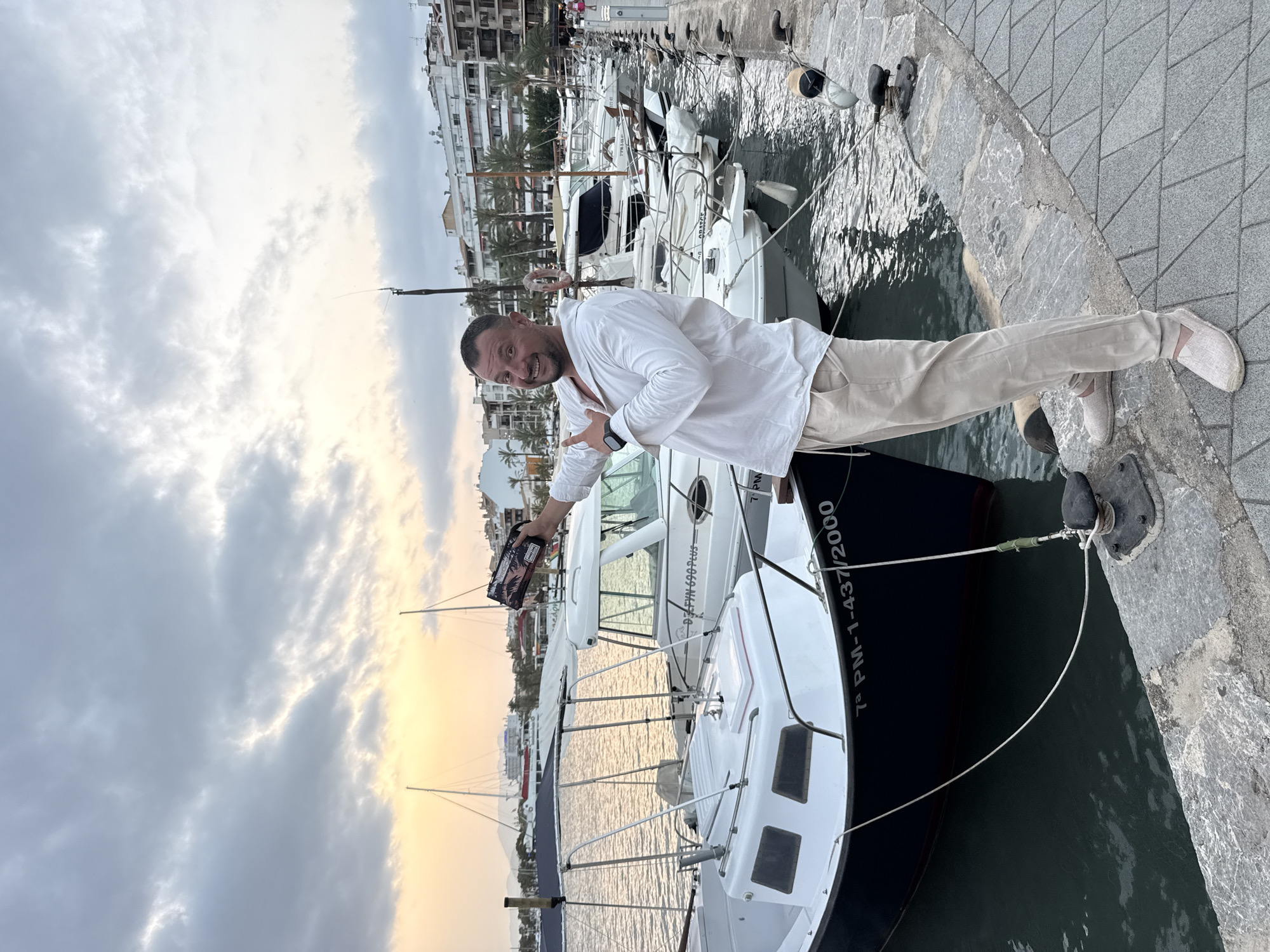 A man in a white linen shirt and beige pants stands on a stone pier in Port D'alcúdia, Spain, holding a small, dark object with a patterned design in his right hand. He is gesturing with his left hand towards the object. Behind him, several white boats are moored in the water, with their masts and rigging visible. In the background, buildings line the waterfront, and palm trees are scattered among them. The sky is cloudy, with a soft glow of sunset on the horizon. The water is calm, reflecting the sky and the boats. The scene appears to be a harbor at dusk.