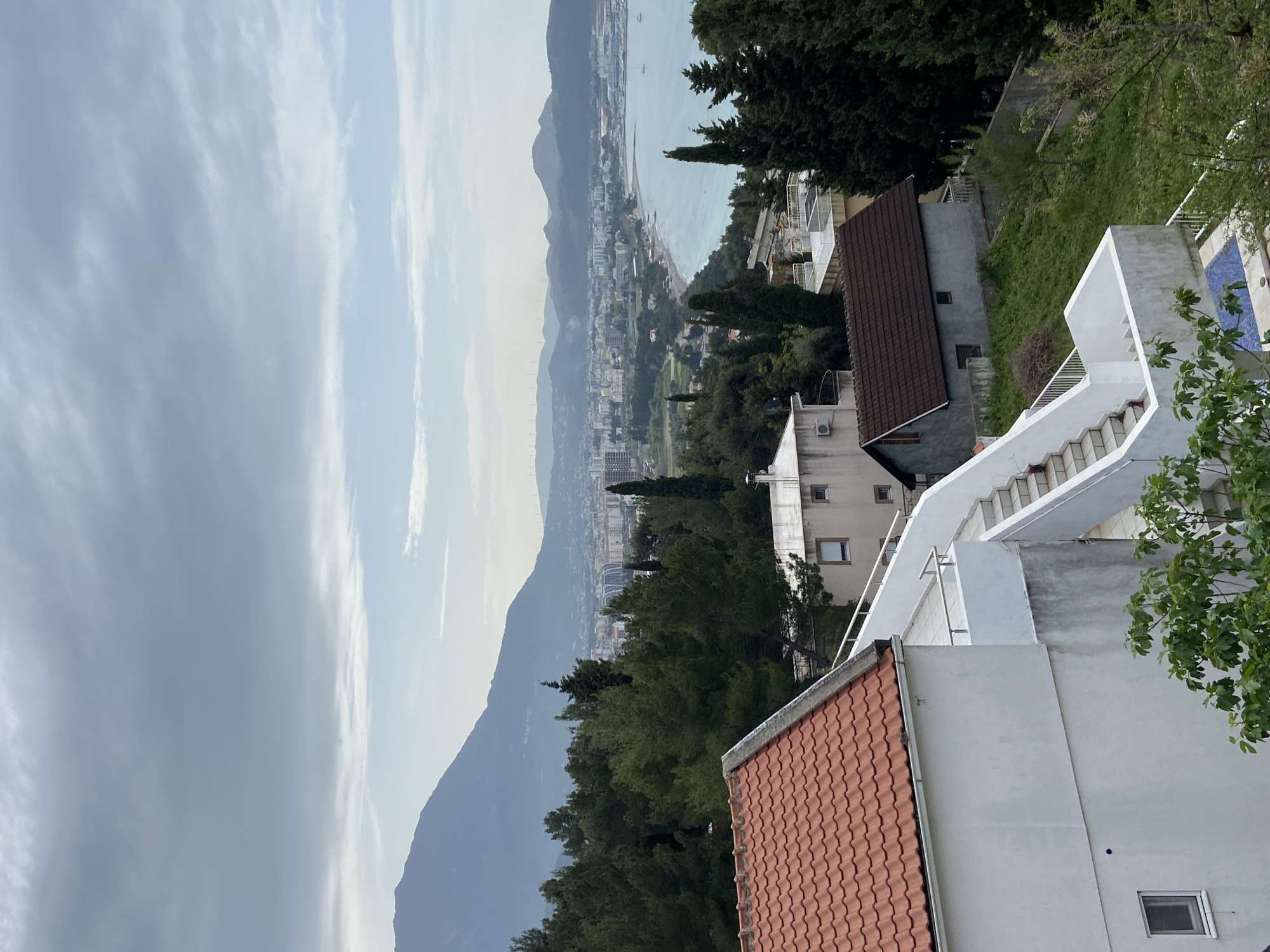 The image depicts a view from Šušanj, Montenegro, showcasing a mix of residential structures and natural scenery. In the foreground, there are white buildings with red-tiled roofs, typical of the region, along with green foliage and trees. The background reveals a coastal city with mountains and the sea visible in the distance, partially obscured by atmospheric haze. The overall impression is one of a built environment integrated within a natural landscape.
