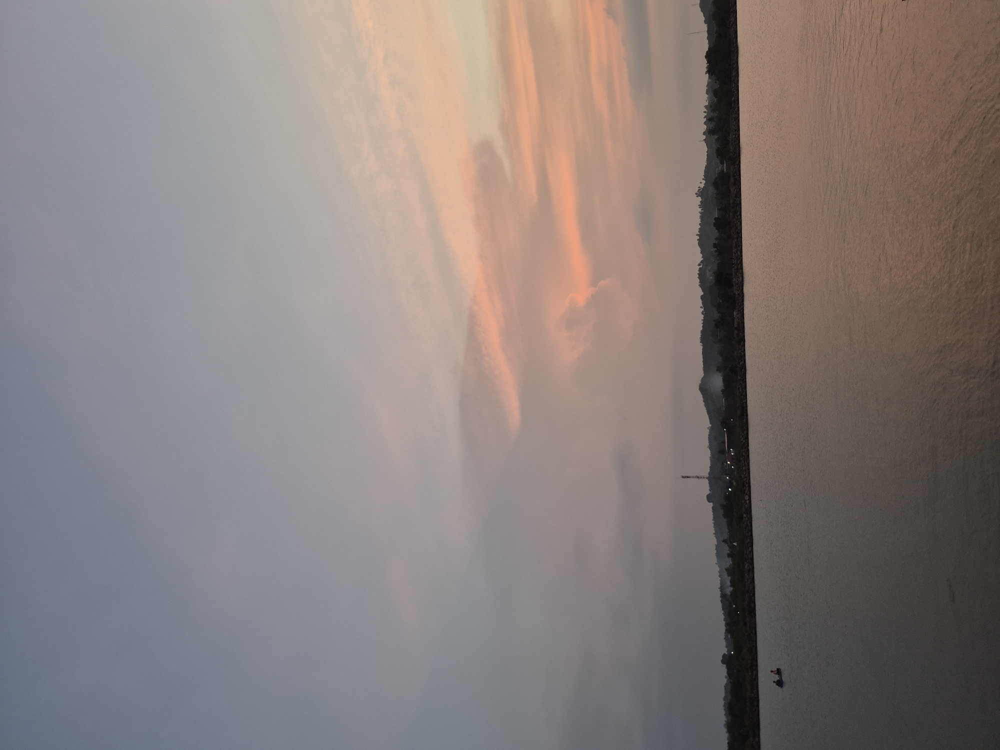 The image depicts a body of water, possibly a lake or sea, with a shoreline visible on the right side. The sky above is a mix of grey and orange hues, indicating either sunrise or sunset. Along the shoreline, there appears to be vegetation and possibly some structures. A small boat is visible in the water near the shore. Given the location is Lhokseumawe, Indonesia, it's likely the water is part of the Malacca Strait or a nearby reservoir. The image overall captures a serene, natural setting with soft lighting.