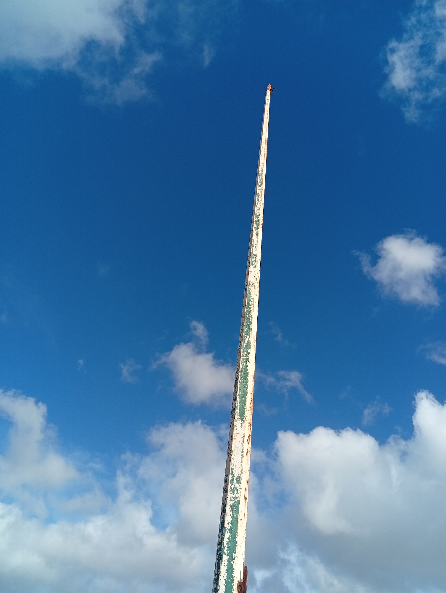 A low-angle, upward shot shows a weathered flagpole against a backdrop of a bright blue sky interspersed with scattered white clouds. The flagpole is painted white with peeling green paint and rust stains, suggesting it is old and in need of maintenance. At the very top of the flagpole, there is a small bird perched. The location is Juan Griego, Venezuela, as indicated in the prompt.