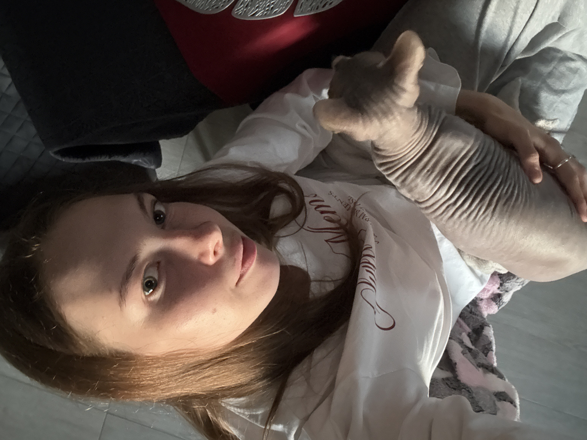 A young woman with brown hair pulled back is holding a hairless cat in her lap. She is wearing a white sweatshirt with a maroon graphic and gray sweatpants. The cat is light brown and has visible wrinkles. The woman is looking at the camera with a slight smile. In the background, a dark blanket and a red object with a decorative pattern are visible. The scene appears to be indoors, possibly in a residential setting. No specific location in Novaya Ivanovka, Russia, can be determined from the image.