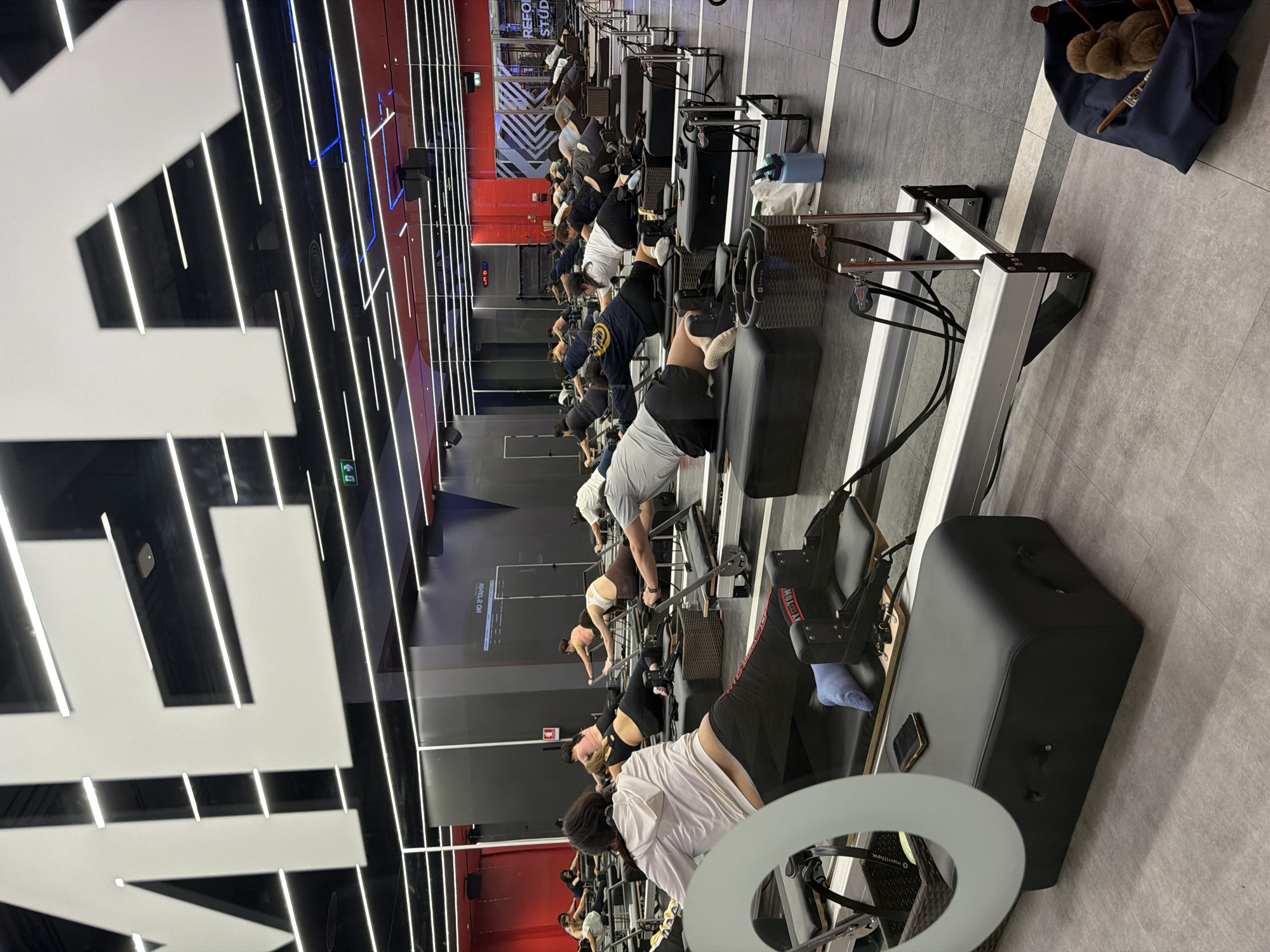 A Lagree fitness class is in session inside a studio in Nong Pa Khrang, Thailand. Approximately 15 people are lined up on reformer machines, engaging in exercises. The studio has a dark ceiling with linear white lights, red and black walls, and a gray floor. A large white graphic is partially visible in the foreground. Several personal items, including a blue bag and a water bottle, are placed on the floor near the machines.