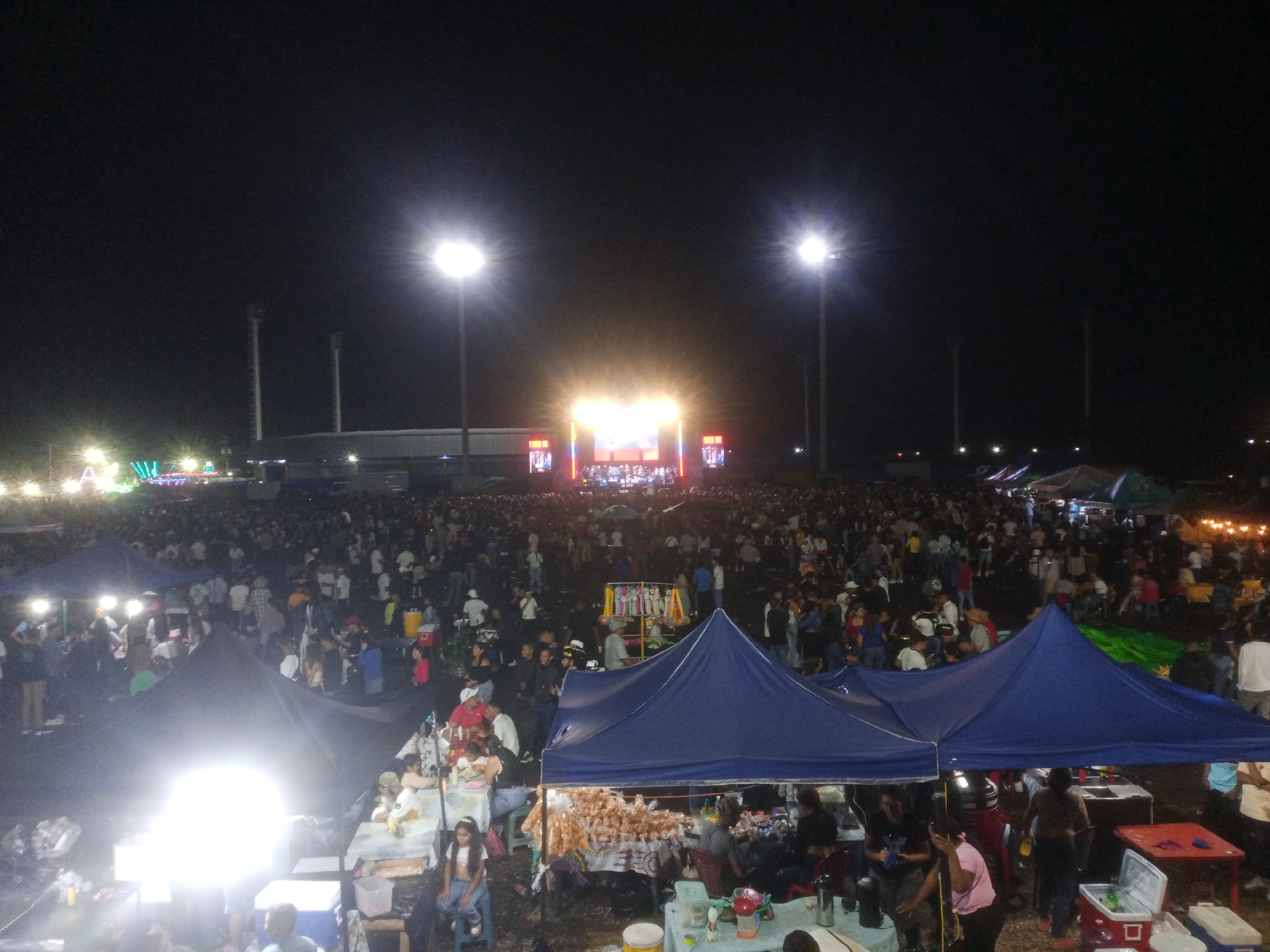 This nighttime aerial shot captures a large, bustling outdoor event in Tinaquillo, Venezuela, likely a concert or festival. The scene is dominated by a vast crowd of people gathered in an open field under a dark sky, illuminated by numerous artificial light sources.
In the mid-ground, a brightly lit stage serves as the central focal point, featuring a performance or presentation. The stage is flanked by tall, stadium-style light poles emitting intense white light. Behind the stage, a large building or stadium structure is dimly visible in the distance.
The area between the foreground and the stage is densely packed with attendees, indicating a significant turnout. In the foreground and extending into the mid-ground, several vendor stalls are set up under various tents, predominantly blue. These stalls are brightly lit, showcasing goods and services. People are visible interacting at these stalls, with some seated at tables under the tents, suggesting food and drink vendors. For example, one blue tent in the foreground shows tables with individuals, including children, and another stall features numerous hanging packaged items, likely snacks or merchandise.
The overall impression is one of a vibrant community gathering taking place after dark, filled with activity and illuminated by the stage lights, vendor lights, and tall floodlights. Due to the distance and lighting, no explicit text or specific logos are clearly discernible on the stage or elsewhere.