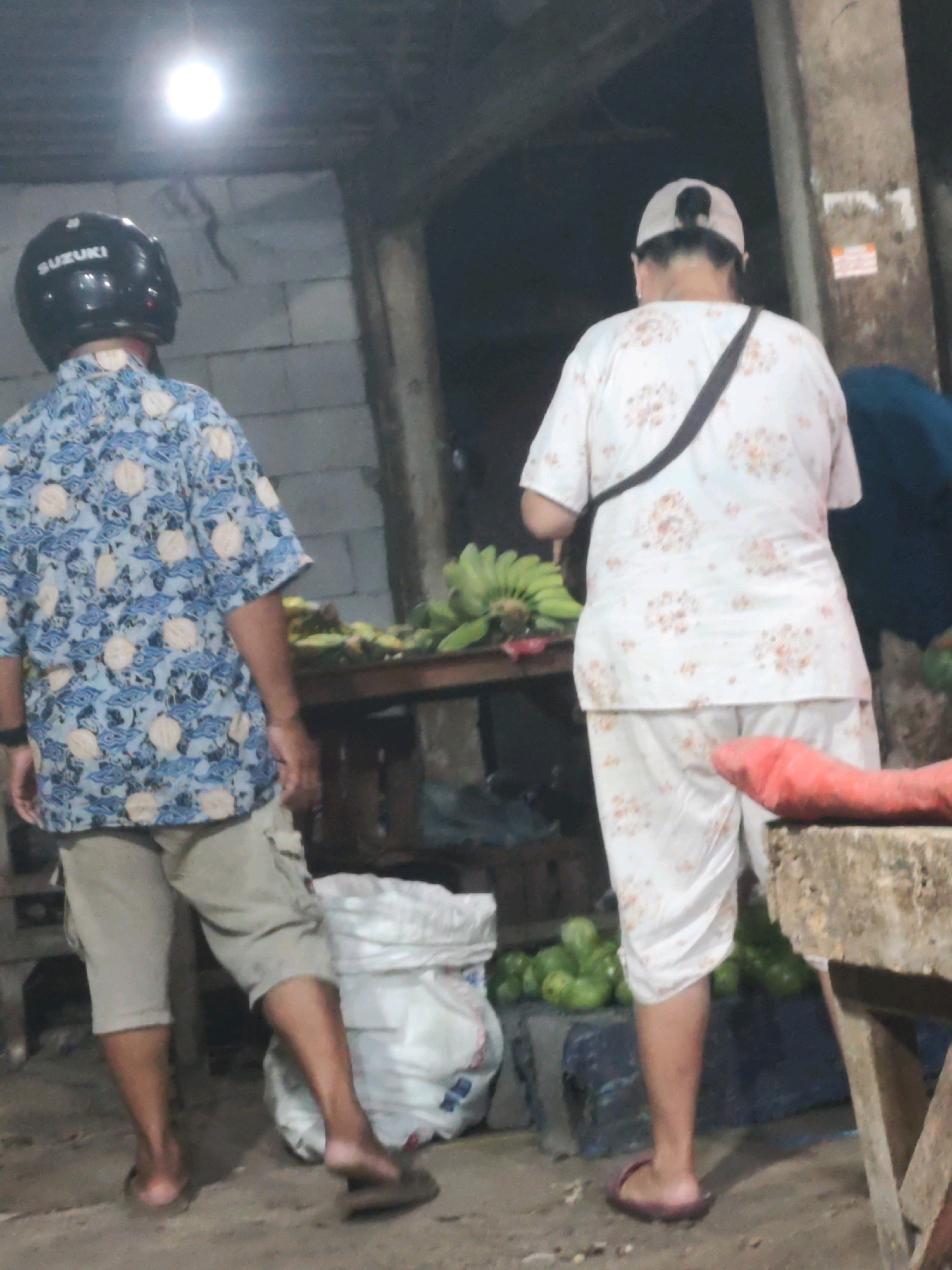 The image depicts a street scene, likely a market, in Karawang, Indonesia. Two individuals are present. The person on the left wears a black helmet with "SUZUKI" branding and a blue patterned short-sleeved shirt with khaki cargo shorts. They are standing with their back to the camera, facing towards a table laden with green bananas. The person on the right, also facing away, wears a white cap and a white short-sleeved shirt with an orange floral pattern, paired with matching white trousers. A dark strap is visible across their shoulder. They are standing near a display of green produce, possibly guavas, and a large white sack. In the foreground, on the right edge of the frame, a red object, possibly a piece of fabric or produce, rests on a wooden structure. The environment appears to be an outdoor market with concrete block walls and a dimly lit ceiling.