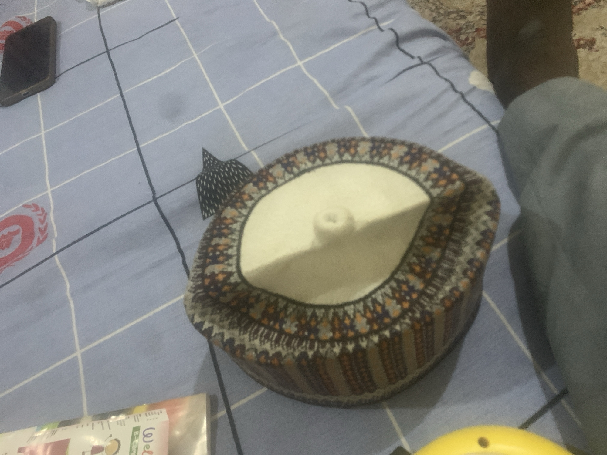 The image shows a close-up, overhead view of a patterned fabric cap, likely a kufi, resting on a blue, checkered surface, possibly a bedsheet or blanket. The cap has intricate geometric patterns in shades of brown, orange, and blue, with a white top. To the left of the cap, a portion of a white plastic package with some text is visible. In the upper right corner of the image, a dark-colored smartphone is placed on the same blue surface. The background is a close-up of the blue checkered fabric, with some shadows and a hint of a rug in the lower right corner. The lighting suggests an indoor environment. There are no people visible in the image. The image does not provide any specific information about the location being in Kuson, Nigeria.