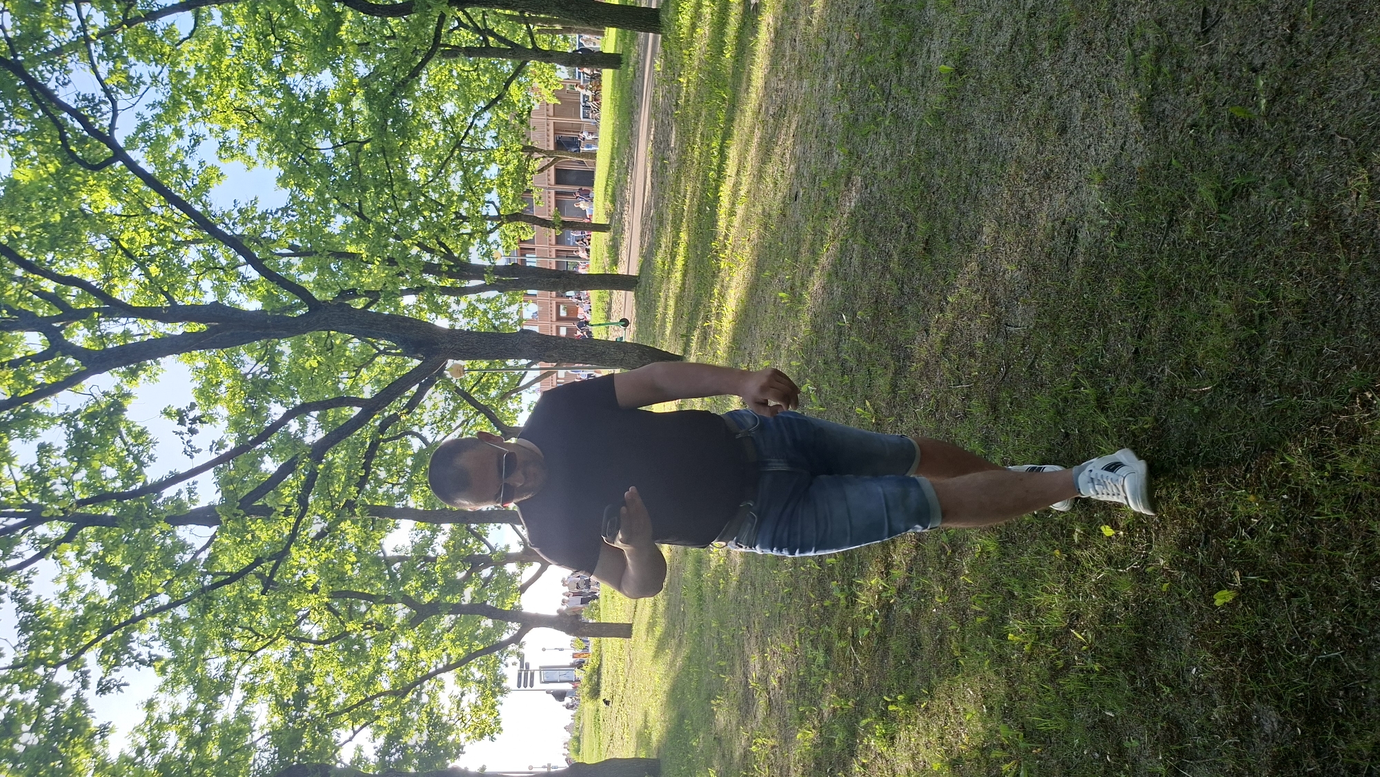 A man in his 40s with dark hair, wearing sunglasses, a black t-shirt, and denim shorts, walks on a grassy area in a park. He is looking down at his mobile phone. In the background, several trees with green foliage line a paved path. Beyond the path, there are buildings and people. The scene appears to be set in the city of Tallinn, Estonia, possibly in a park like Kadriorg Park or a similar urban green space. The sun is shining, casting shadows on the grass.