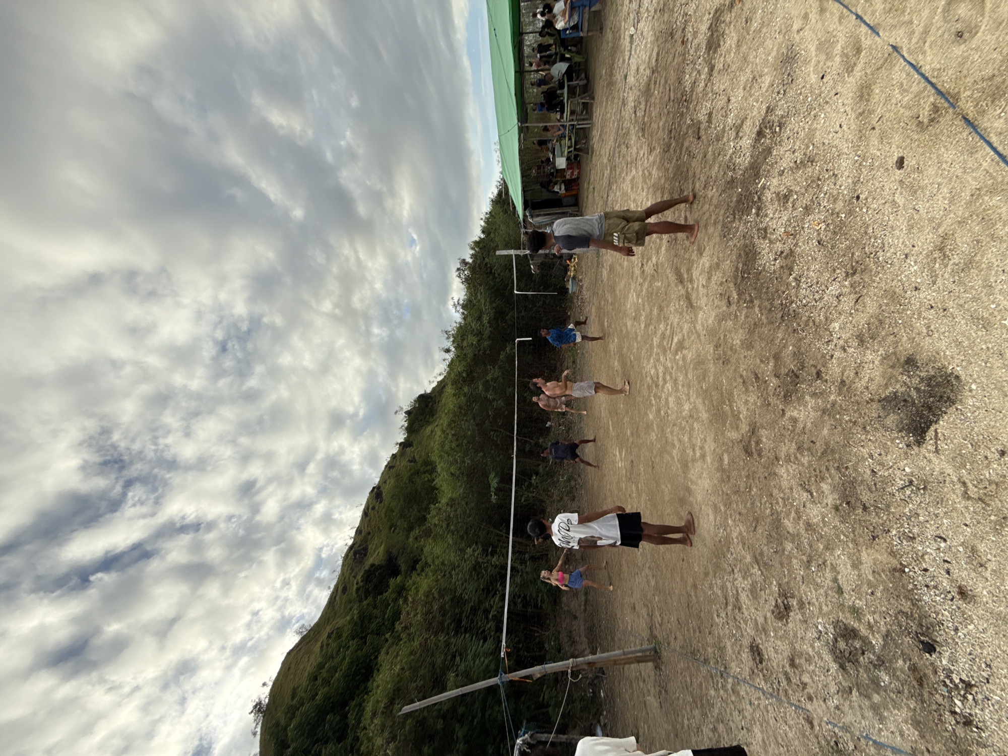 The image shows a beach volleyball game in Rinca Village, Indonesia. A volleyball net is visible with several people standing on the sand playing or preparing to play. A forested hill rises in the background to the left. Behind the volleyball court, further in the background, is a covered area with people seated and watching. The sky is cloudy. The beach sand is light-colored.