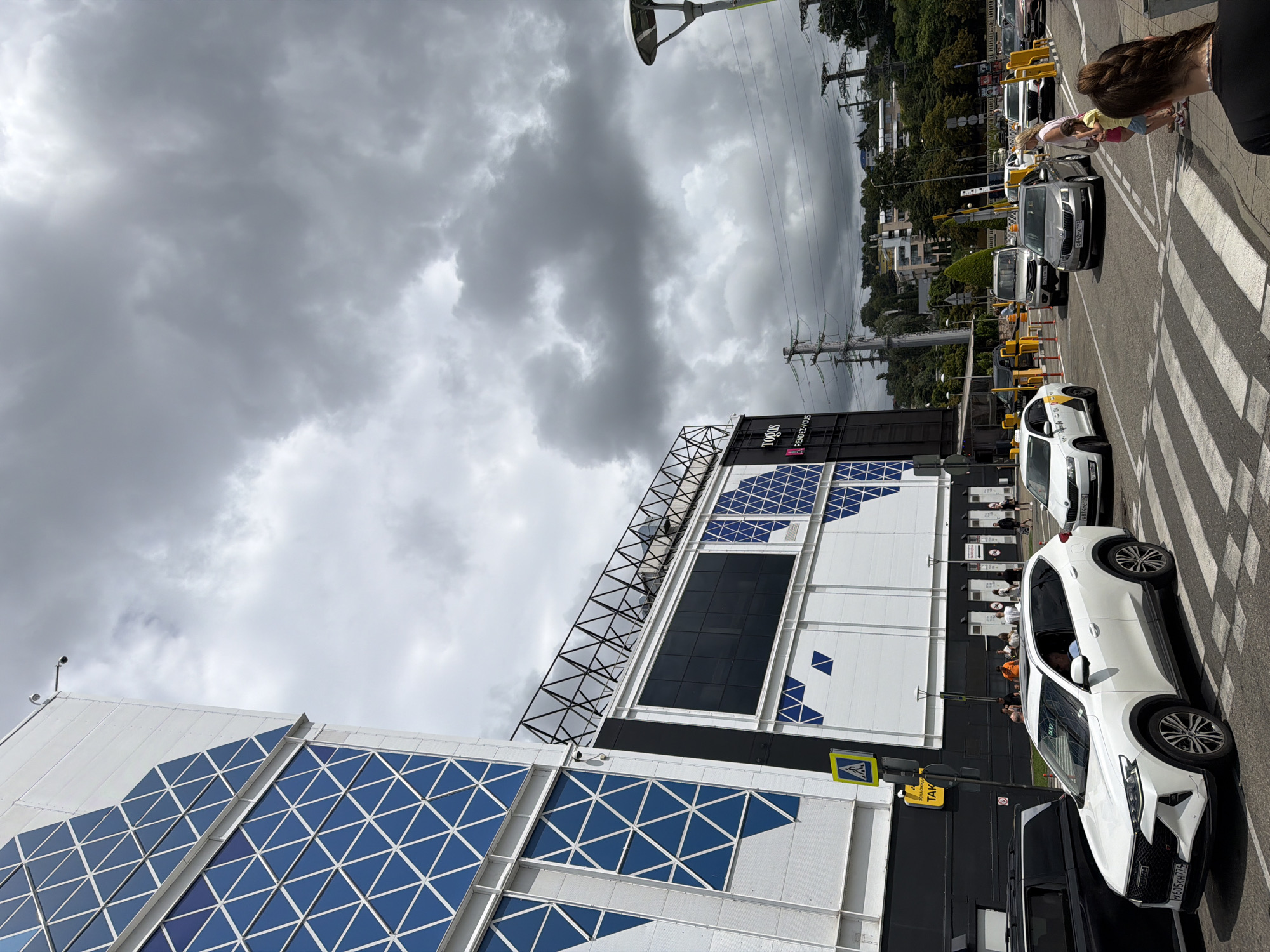 The image captures a daytime scene on a cloudy day in Sochi, Russia, near a large modern building complex. In the foreground, a white SUV is stopped at a pedestrian crossing, with its front license plate visible. To its left, a black vehicle is partially visible. Further down the road, several other cars are in motion or stopped, including a silver Skoda and a white sedan. People are seen walking on the sidewalk near the building. The building itself is large and contemporary, with a facade featuring geometric blue patterns and large dark windows. A yellow sign with a pedestrian symbol is mounted on a pole near the building. In the background, power lines and trees are visible, and the sky is filled with heavy, dark clouds, suggesting an impending storm. The location is identified by signage on the building as "TODOS RENDEZ-VOUS".