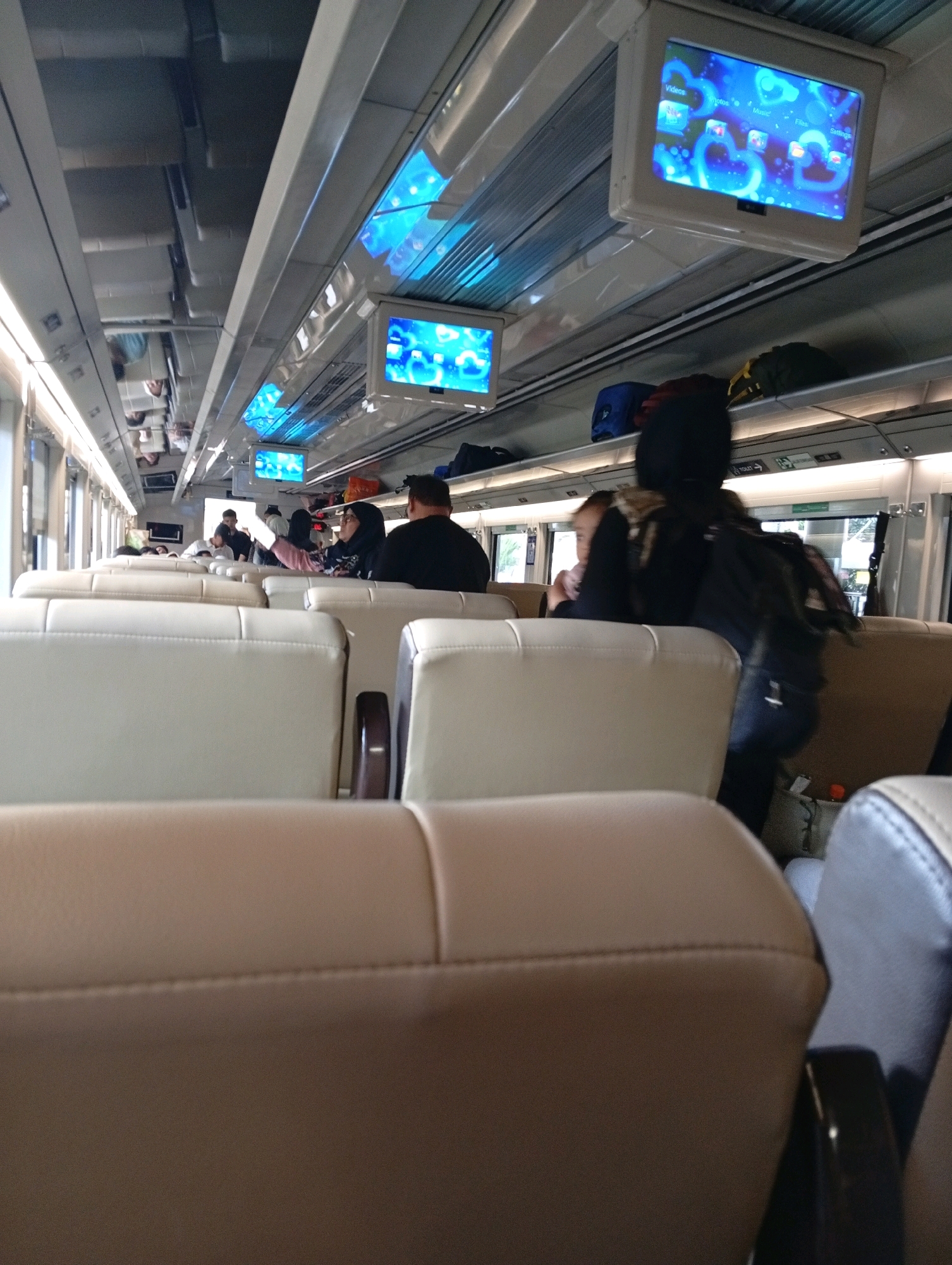 The image depicts the interior of a train carriage, likely on a long-distance route within Indonesia, possibly in Yogyakarta. The perspective is from a seated passenger looking forward, showing rows of beige and light brown upholstered seats. The ceiling of the carriage is visible, with fluorescent lighting and overhead luggage racks. Two small screens are mounted on the ceiling, displaying a blue interface with icons. Several passengers are visible in the background, some seated and others standing. One passenger in the foreground, seen from the back, is wearing a black jacket and dark jeans. Another passenger, partially visible to the left, is wearing glasses and a headscarf. Luggage, including a blue bag and a dark green bag, is stored on the overhead racks. The overall environment suggests travel, with a focus on the seating arrangement and in-carriage amenities of the train.