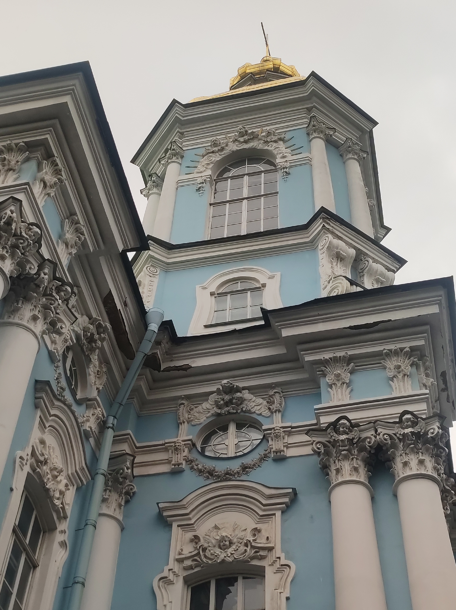 This image depicts a low-angle view of a building with ornate blue and white Baroque architecture. The building features elaborate white stucco decorations, including scrolls, floral motifs, and sculpted cherubs adorning the cornices and columns. There are arched windows with multi-paned glass, and a golden dome with a cross on top is visible in the upper part of the frame. The sky is overcast and a light blue gutter runs vertically down the side of the building. This is the Smolny Cathedral, located in Saint Petersburg, Russia.
