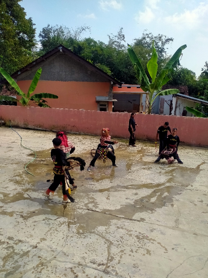 An outdoor scene in Tawangsari, Indonesia, captures six individuals on a wet concrete surface under a clear sky. In the foreground, three subjects engage in a traditional performance. The leftmost subject, a young male in a black and gold garment with white shoes, steps forward holding a decorated horse-like prop. To his right, another individual with a red head covering and patterned attire is partially obscured. Further right, a performer wearing a brown mask and patterned clothing is in a wide stance with extended arms. A green garden hose is coiled on the wet concrete to the left. In the background, three additional individuals, dressed in black, are positioned near a low orange wall. One individual on the right holds a whip-like object. Two others are standing and kneeling near a yellow rod. Behind the wall, an orange house with a sloped roof and a partially visible structure are present, framed by dense green foliage and trees.