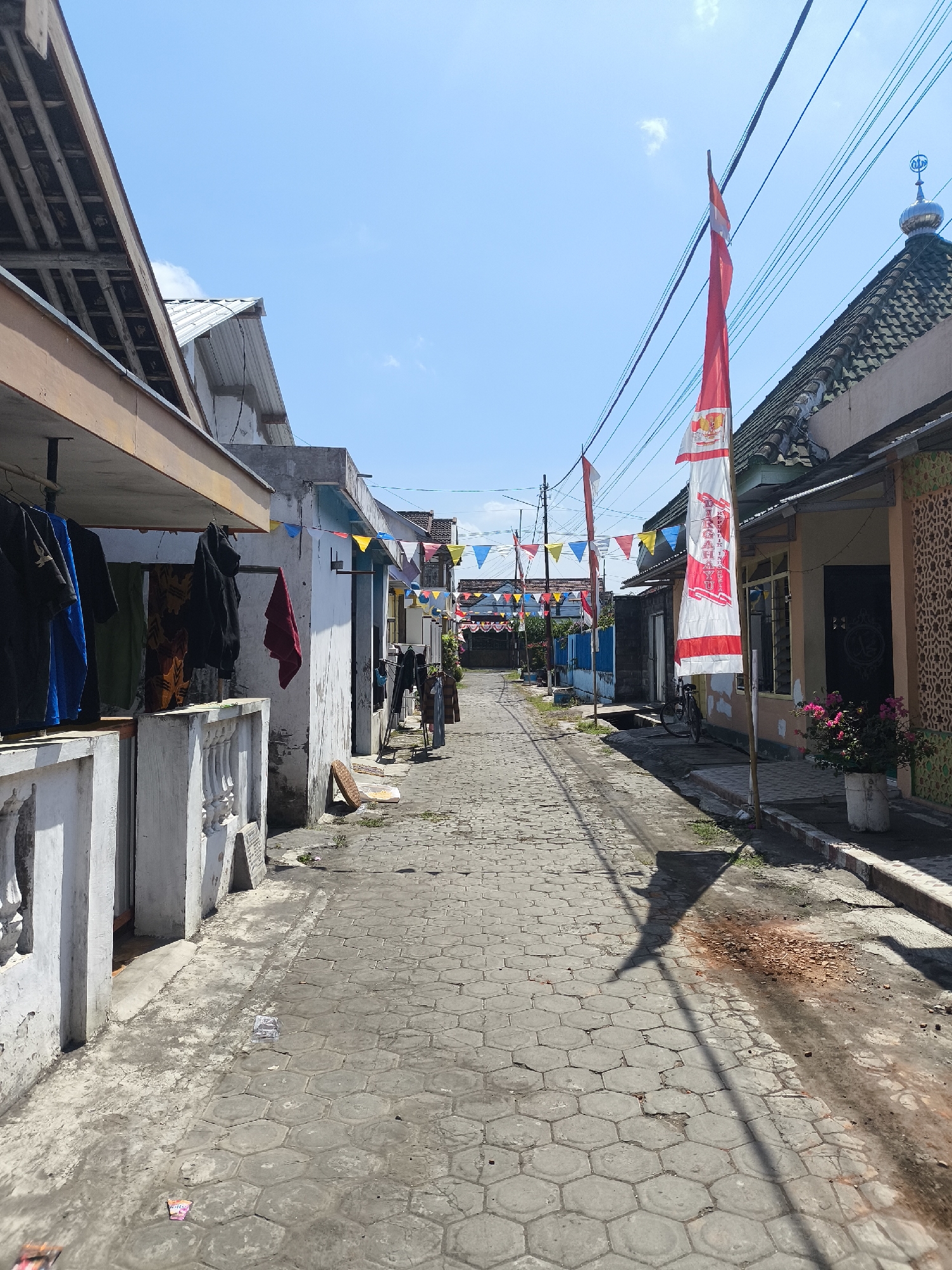 The image shows a narrow, paved street in Lumajang, Indonesia. The street is lined with houses and decorated with Indonesian flags and colorful triangular flags, suggesting a celebratory occasion. On the left side of the street, clothes are hanging on a clothesline in front of a house. On the right side, a bicycle is parked near a house with a tiled roof and a dome-shaped structure on top, possibly a mosque. The sky is clear and blue. The ground is paved with hexagonal stones, and there is some debris on the side of the road. The overall scene depicts a residential area in Lumajang during a festive event.