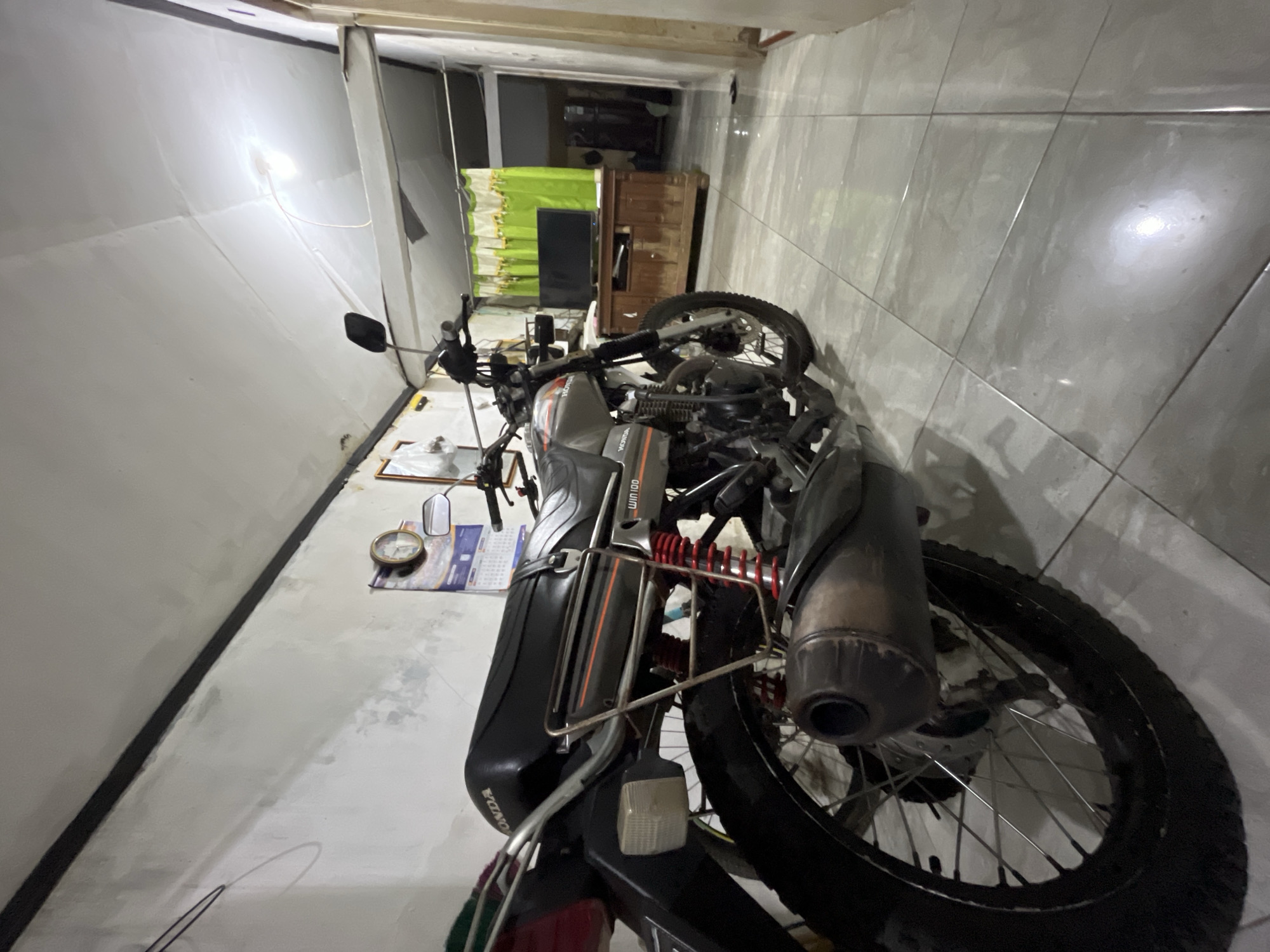 The image depicts the interior of a dwelling in Pekalongan, Indonesia. A Honda Win 100 motorcycle is parked indoors, occupying a significant portion of the foreground and midground. The motorcycle is gray with orange and black detailing. The interior features a tiled floor, white walls, and a white ceiling with visible light fixtures and some signs of wear. In the background, a wooden cabinet with a television and green curtains are visible. A clock and a calendar are mounted on the wall. The scene appears to be a residential space, possibly a garage or an area within a house used for storage. There are no people visible in the image.