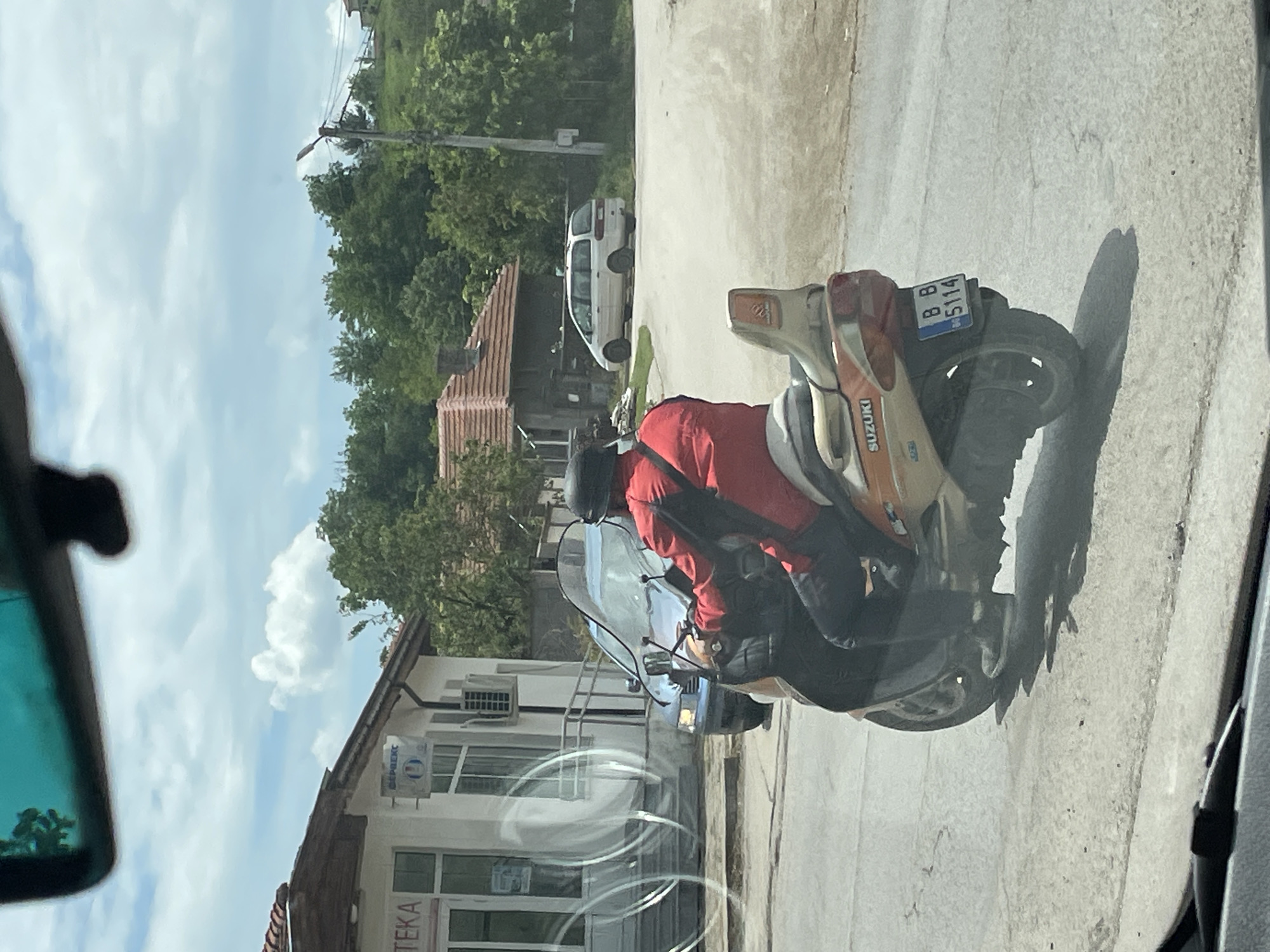 A photo taken from inside a car shows a man on a Suzuki motorcycle with license plate BB 5114 in Tsar Kaloyan, Bulgaria. The man wears a red jacket and helmet. The motorcycle is parked on the side of the road in front of a building with the word "TEKA" visible, and a white van is parked further down the street. The environment appears to be a residential area.