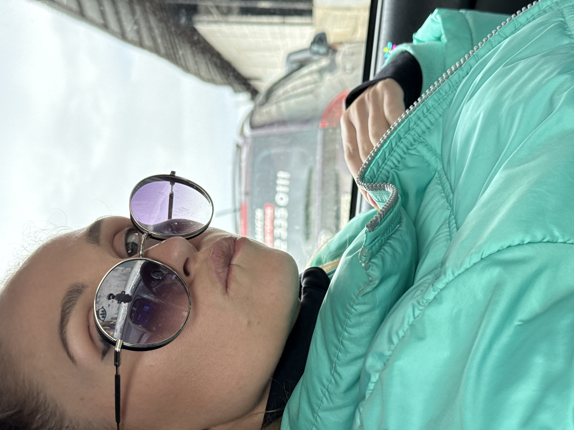 An image, rotated 90 degrees counter-clockwise, depicts a person from the chest up, seemingly looking forward or slightly upwards. The individual has light skin, dark eyebrows, and wears large, round sunglasses with dark frames. The left lens (viewer's left) appears purple-tinted, while the right lens reflects the outside environment, showing a dark, indistinct object and some shapes. The person's lips are slightly parted.
They are wearing a mint green puffy jacket with a silver zipper visible along the collar and front, and a black long-sleeved garment underneath. The background is an out-of-focus view through a window, suggesting the person is inside a vehicle. The sky outside is overcast and light grey.
Visible through the window, behind the person, is a blurry street scene. A large vehicle, possibly a bus or truck, is prominent, displaying partial text in white: "1110 СЕЕ" (likely Cyrillic "1110 CEE") and a phone number "333 0111". These details align with the provided location context of Chaikovsky, Russia. The overall impression is a casual, relaxed moment inside a vehicle during daytime.
