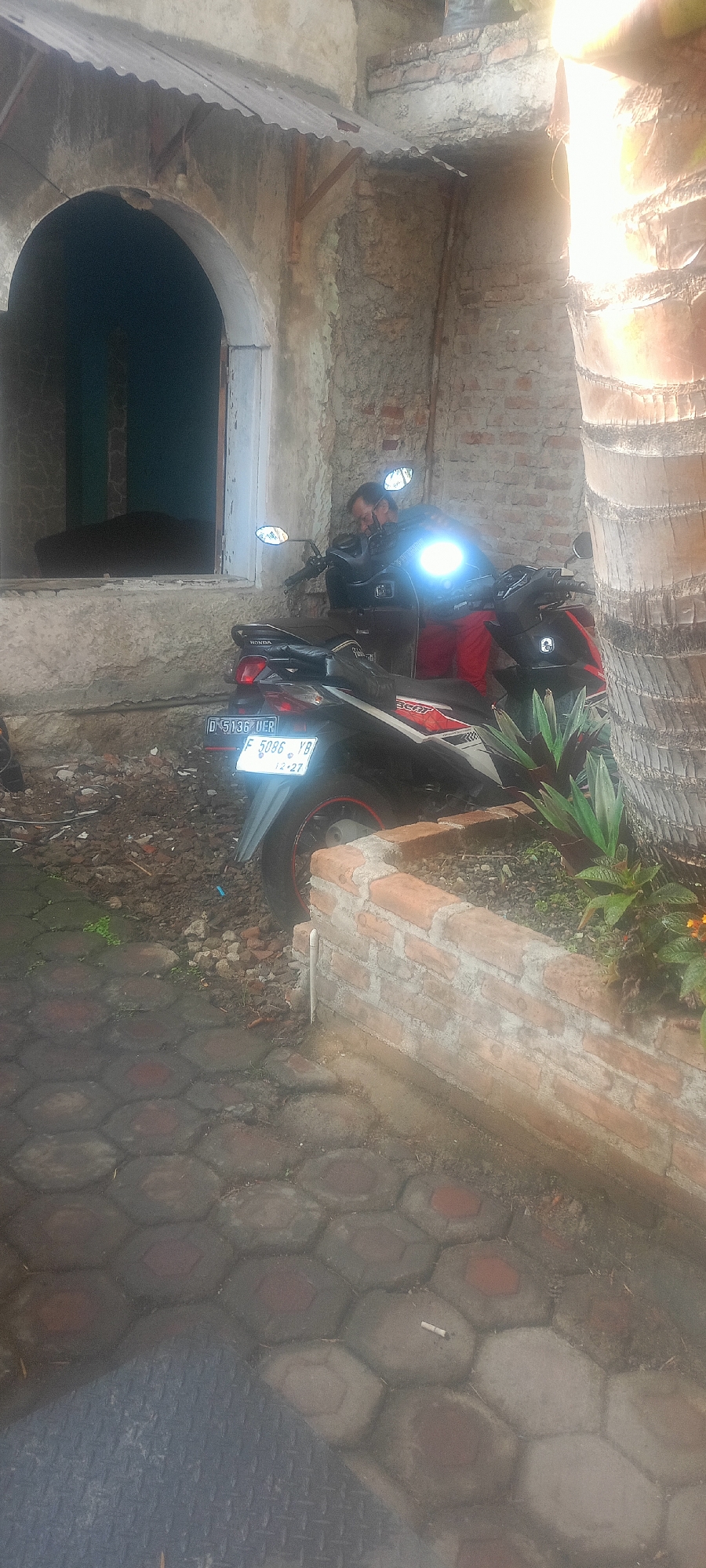 In an outdoor scene in Mekar Rahayu, Indonesia, a man wearing glasses and a dark jacket is leaning over a black motorcycle. The motorcycle has a bright headlight illuminated and is parked on a cobblestone pathway. The man is partially obscured by the motorcycle and the bright light. Behind the motorcycle, there is a wall constructed of bricks and concrete. To the left of the motorcycle, an arched doorway is visible, leading into a dark interior. To the right of the motorcycle, a palm tree trunk and some green foliage are present, with a low brick planter in the foreground. The ground is covered in a mixture of dirt, debris, and the hexagonal cobblestones. A cigarette butt is visible on the cobblestones.
