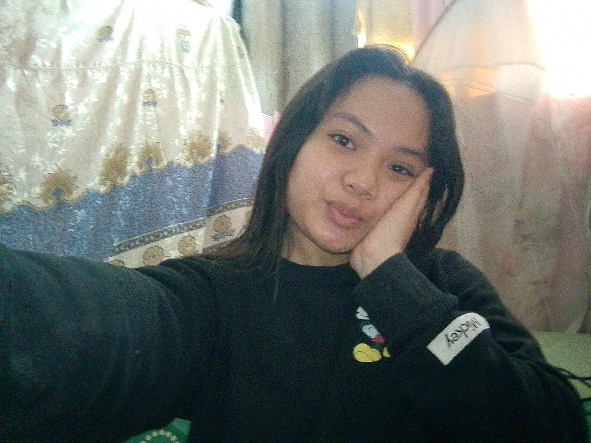 A young woman with dark hair and brown eyes is taking a selfie. She is wearing a black long-sleeved shirt with a Mickey Mouse graphic on the left sleeve and a white tag with "Mickey" written on it. Her right hand is resting on her cheek, and she is looking at the camera with a slight smile. She is indoors, with a patterned curtain and what appears to be a bed in the background. The lighting suggests it is daytime.