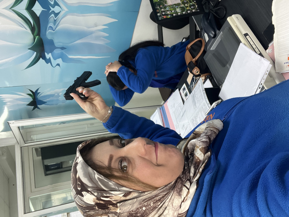 An indoor office scene in Meknes, Morocco, captured from a close-up perspective. In the foreground, a female subject, wearing a beige patterned hijab and a blue zippered fleece top, smiles directly at the camera. Her right hand is raised, holding a black object resembling a compact submachine gun or toy replica. Her immediate workspace includes stacks of documents, a white printer/scanner, and a black corded telephone. In the mid-ground, a second female subject, also in a blue top, is seated at a desk with her hands placed on top of her head, facing away from the camera. Her desk displays a computer monitor with a vibrant desktop background and a brown and black handbag. The background features a light blue wall adorned with a mural depicting white lotus-like flowers with green bases, reflected as if in water. A glass door or partition is visible on the left side of the frame, showing reflections of an adjacent room.