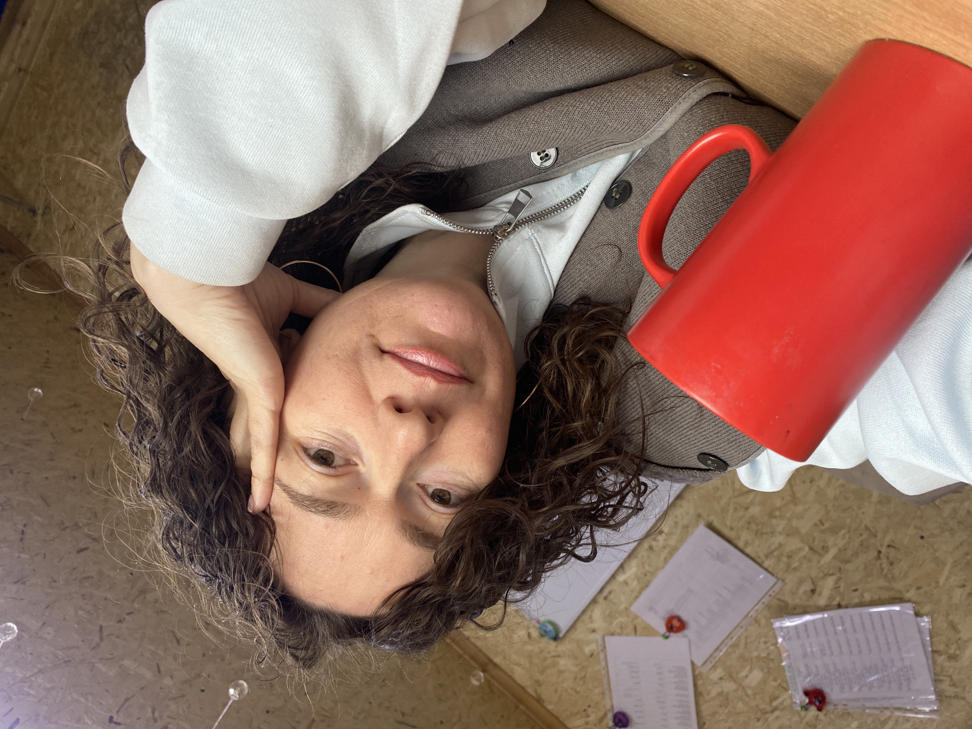 A woman with curly brown hair is lying down, her head propped up by her left hand. She is wearing a white sweatshirt and a brown cardigan. A large red mug is positioned to her right. Several sheets of paper are visible on the surface below, held in place with colorful push pins. The location is possibly indoors, on a wooden surface. There is no evidence to confirm the location in Ol'khovskoye, Russia.