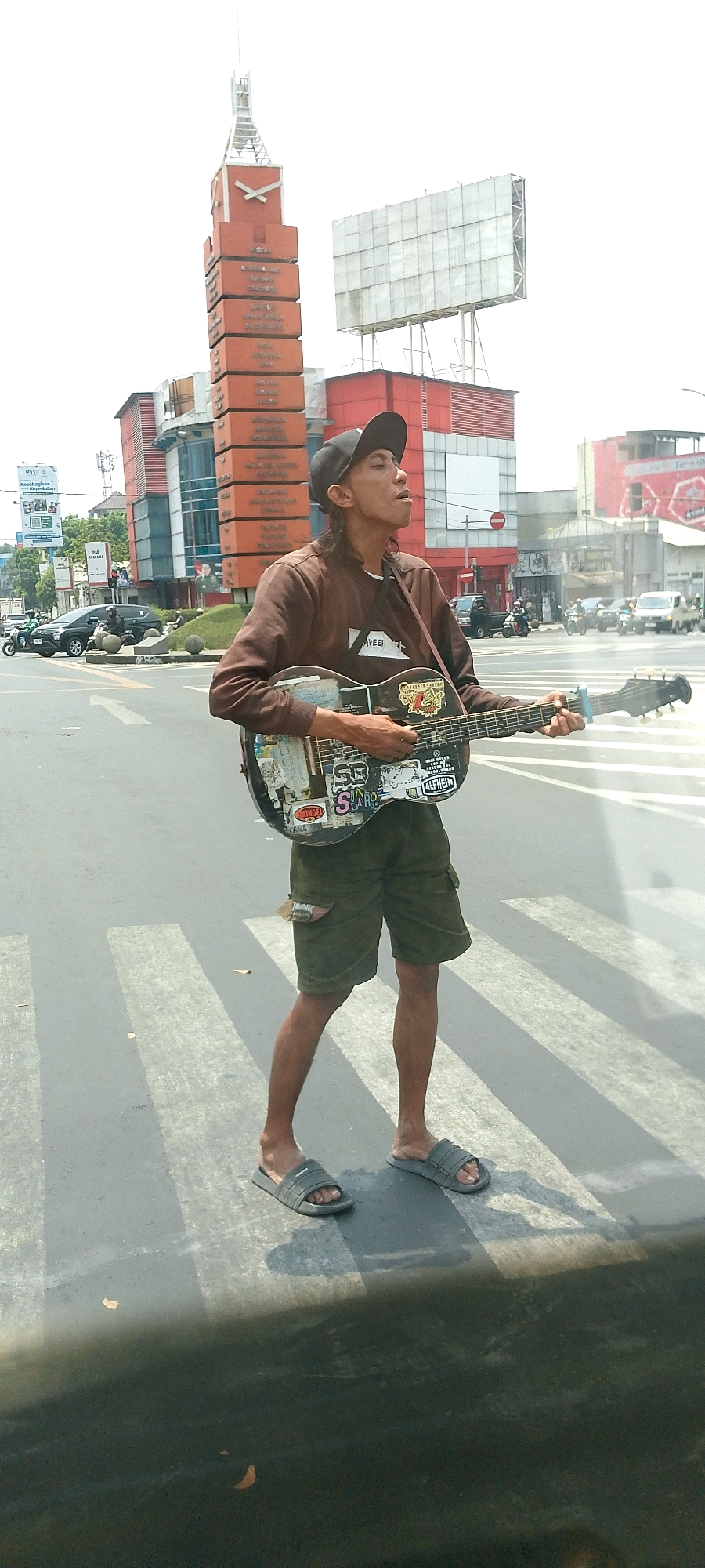 An individual stands on a pedestrian crossing in what appears to be a city street in Bandung City, Indonesia. The person is holding and playing an acoustic guitar, which is heavily decorated with stickers. They are wearing a brown long-sleeved shirt, green cargo shorts, a black baseball cap, and grey slide sandals. The individual is facing slightly to the right and their mouth is open, suggesting they are singing or speaking. In the background, a tall, orange, tower-like structure with writing on it is visible, along with buildings and traffic, including cars and motorcycles. The sky is bright and overcast. The pedestrian crossing has white stripes on a grey asphalt surface.
