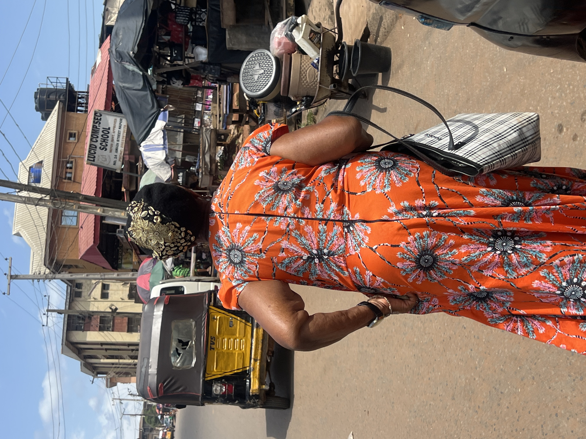 A slightly high-angle outdoor shot shows a woman wearing a vibrant orange floral dress. She has a black and white checkered purse slung over her shoulder and is standing next to a three-wheeled vehicle labeled "TVS."
In the background, there are several buildings and structures, including a sign that reads "LUCID COMP.SEC SCHOOL," indicating the location is likely near this school. The street is lined with miscellaneous objects and tents. Utility poles with wires are also visible against a clear sky. The overall setting is that of a bustling urban area.