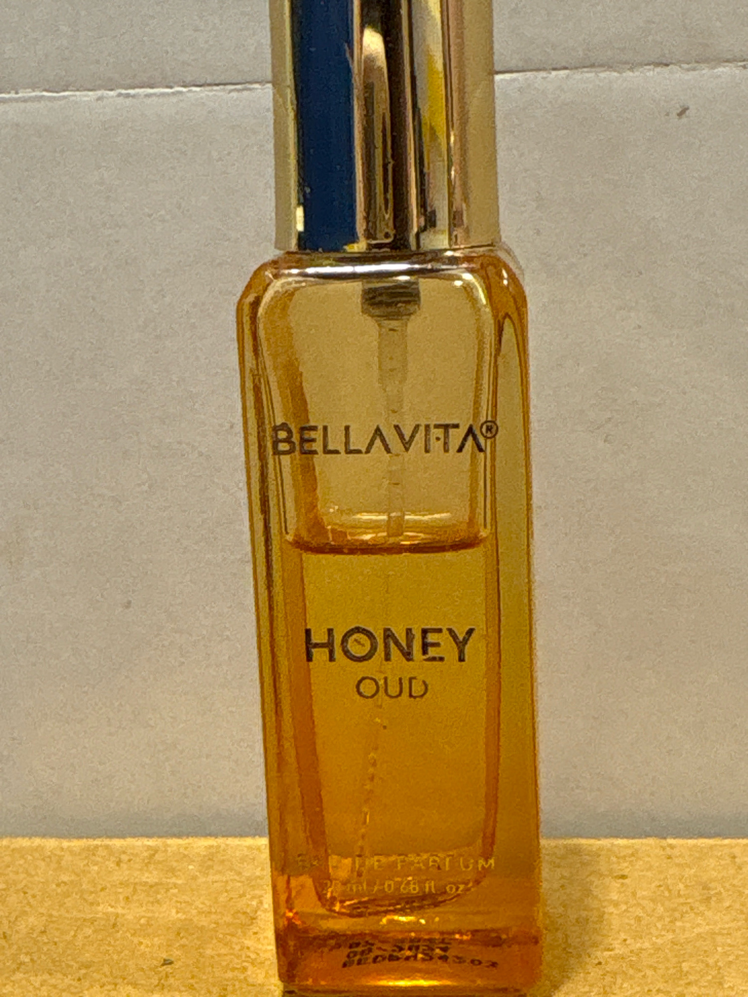 A close-up, eye-level, medium shot captures a bottle of "BELLAVITA HONEY OUD" perfume standing upright on a textured, light brown surface. The bottle is made of amber-colored glass and has a metallic gold cap and collar. The label is black and reads "BELLAVITA" at the top, followed by "HONEY" and "OUD" below. The liquid inside the bottle is a rich golden-yellow. The background is a textured, light grey surface. The lighting is bright, casting reflections on the bottle's surface. There are no people or animals visible in the image, and no specific location in Bhuj, India, can be identified from the scene.