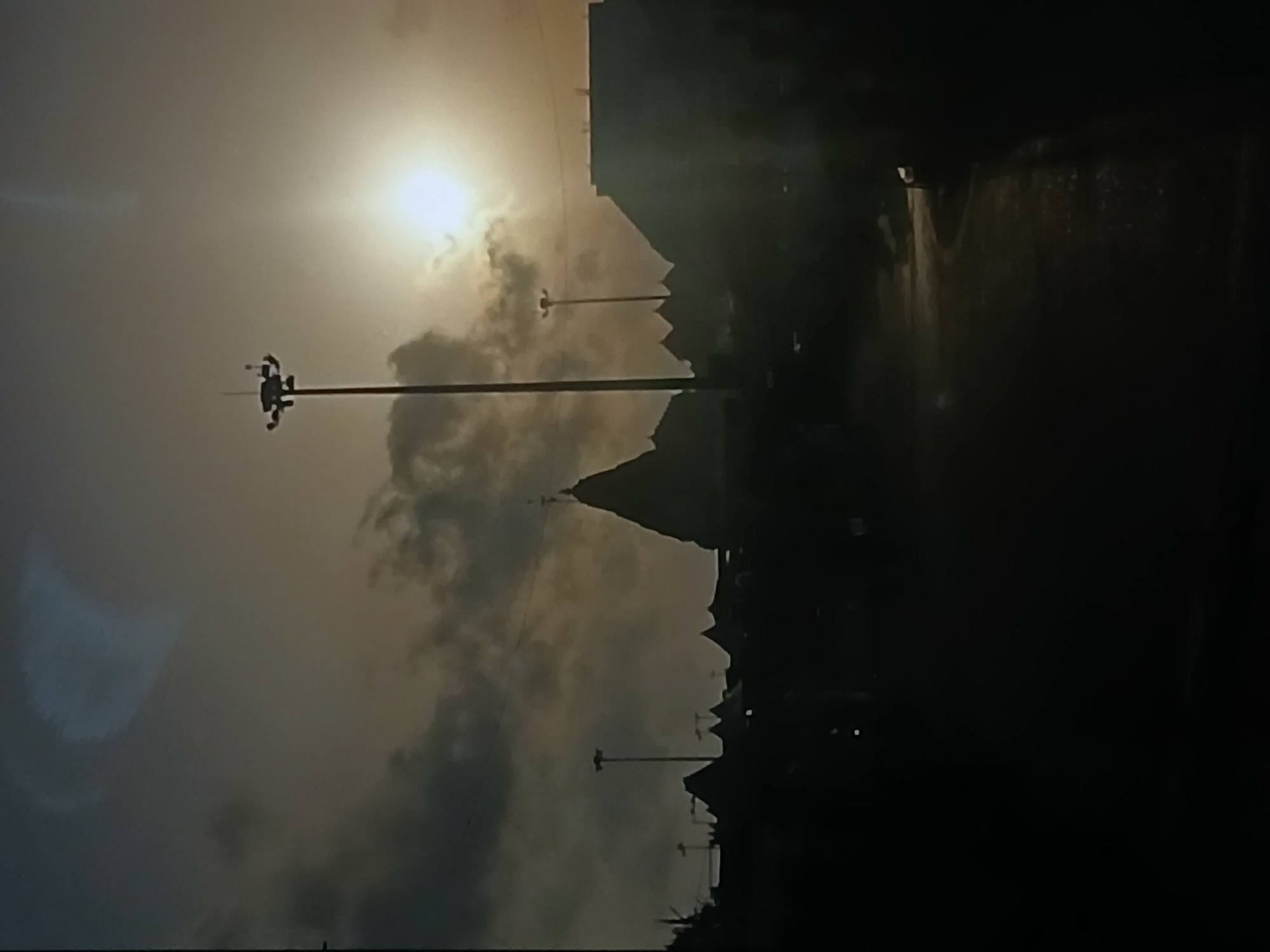 The image presents a silhouetted view of a street scene in Bengaluru, India, likely captured during dawn or dusk. The sun is visible behind clouds, creating a bright haze. A tall lighting pole stands prominently on the left, and there is another smaller one on the right. The buildings are in dark silhouette. The road appears wet, possibly from recent rain.
