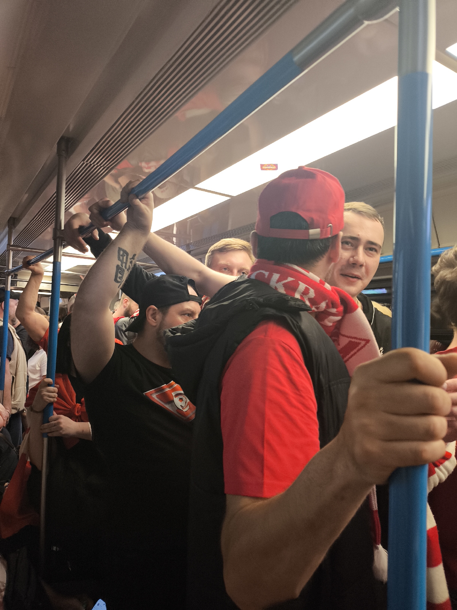 The image shows a crowded subway car, likely in the city of Unknown, Unknown. Several people are visible, primarily in the foreground, holding onto the overhead bars. One person is wearing a red cap and a red and white scarf, while another has a tattoo on their arm. The interior of the subway car has fluorescent lighting. The overall atmosphere appears congested and lively.