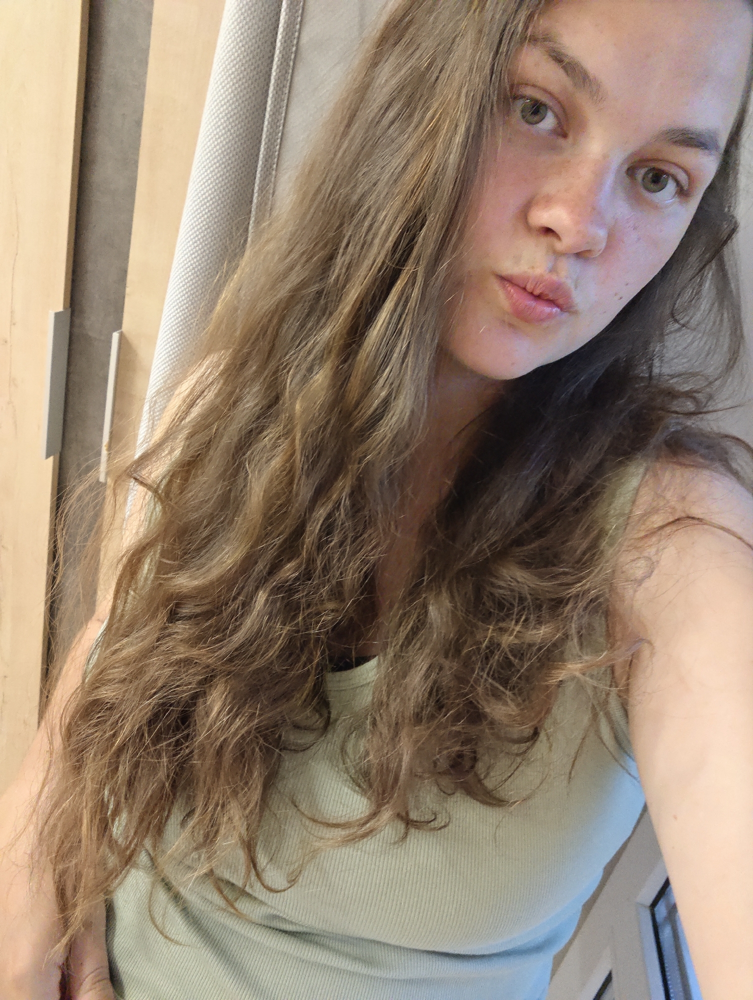 A young woman with long, wavy brown hair and freckles is posing for a selfie. She has green eyes and is puckering her lips. She is wearing a light green ribbed tank top. The background includes a light brown wooden door, a gray textured panel, and a white window frame. There is no information to determine the location in the city of Chaikovsky, Russia.