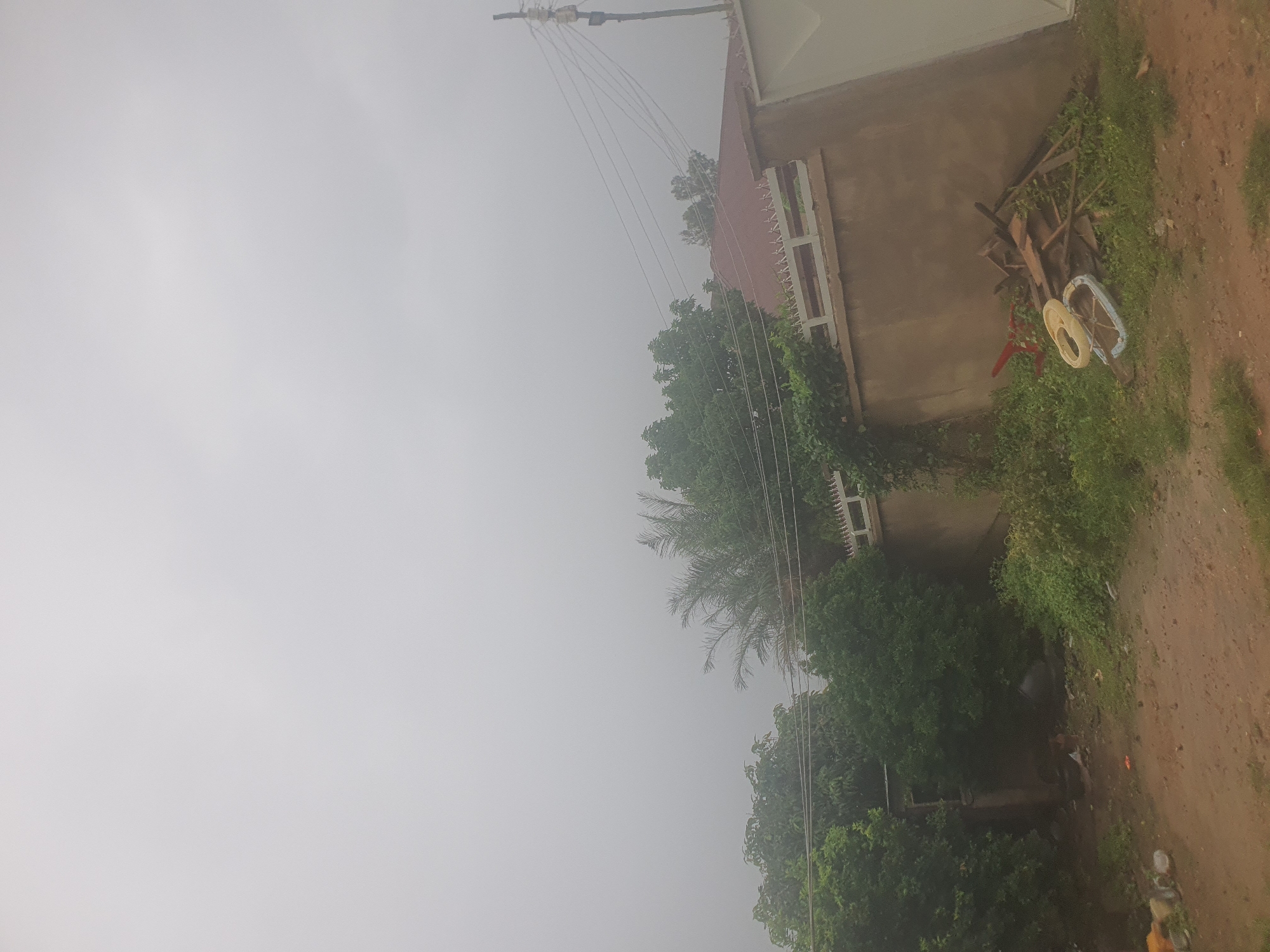 The image depicts a cloudy sky over a suburban area, possibly in Jos, Nigeria. In the foreground, a dirt path leads towards a wall with a tiled roof and a decorative white fence. To the left of the wall, lush green trees and bushes are visible, with some debris and discarded items scattered on the ground. Among the debris, a red object resembling part of a toy, a yellow ring, and a blue object are present. Further to the left, more dense foliage and a corner of a building can be seen. Overhead, power lines stretch across the frame, supported by a utility pole. The overall atmosphere is overcast, suggesting it might be a hazy or rainy day.