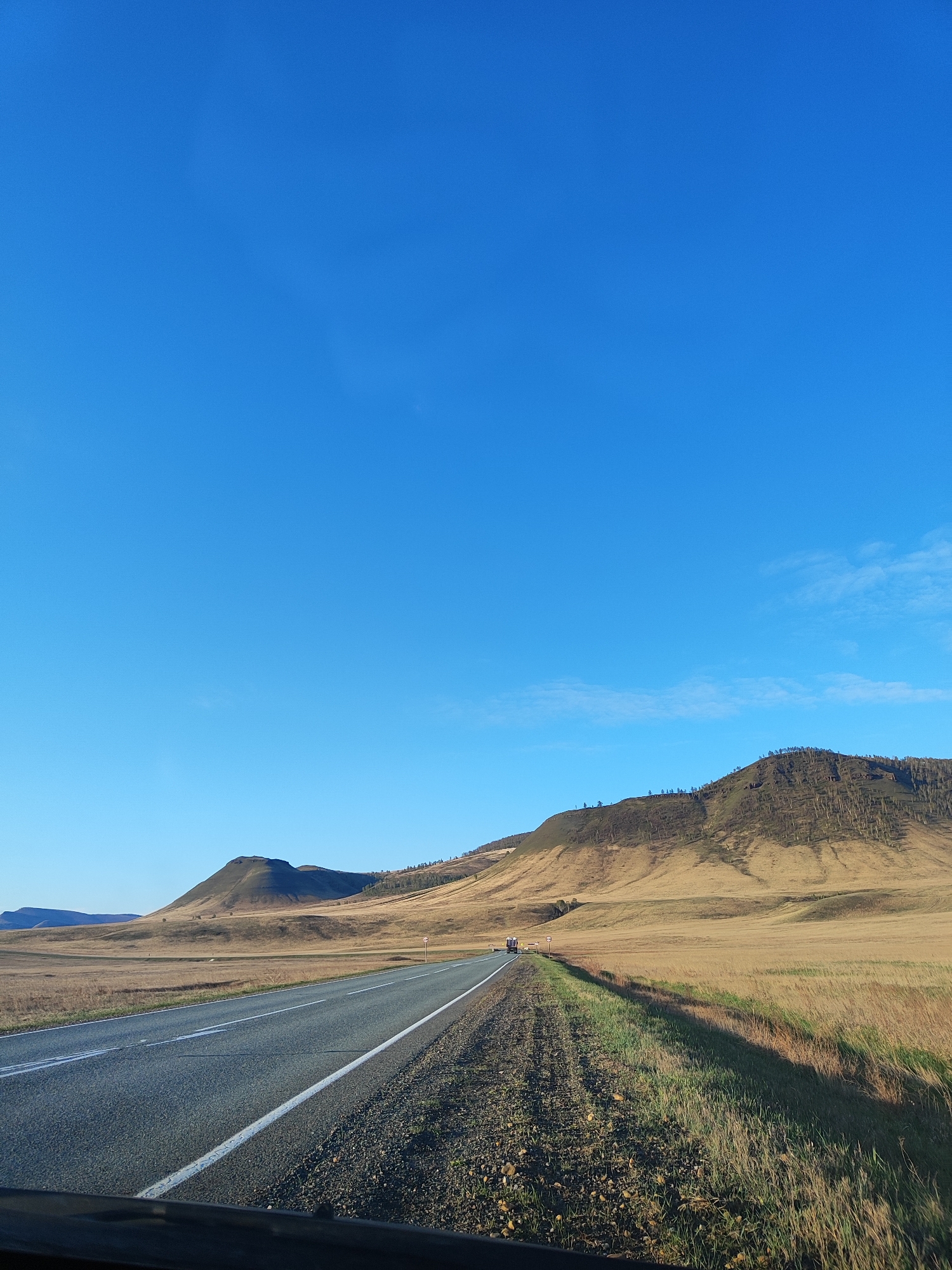 A landscape photograph depicts a straight asphalt road leading into the distance under a clear blue sky with minimal cloud cover. The road is marked with a single white line on either side. To the left and right of the road are dry, golden fields that appear to be grasslands or meadows.
In the background, there are low-lying mountains or hills covered in dry vegetation and sparse trees. The lighting suggests it is either early morning or late afternoon, casting shadows on the landscape. A vehicle is visible on the road in the distance. The perspective suggests the photo was taken from inside a vehicle, likely a car.