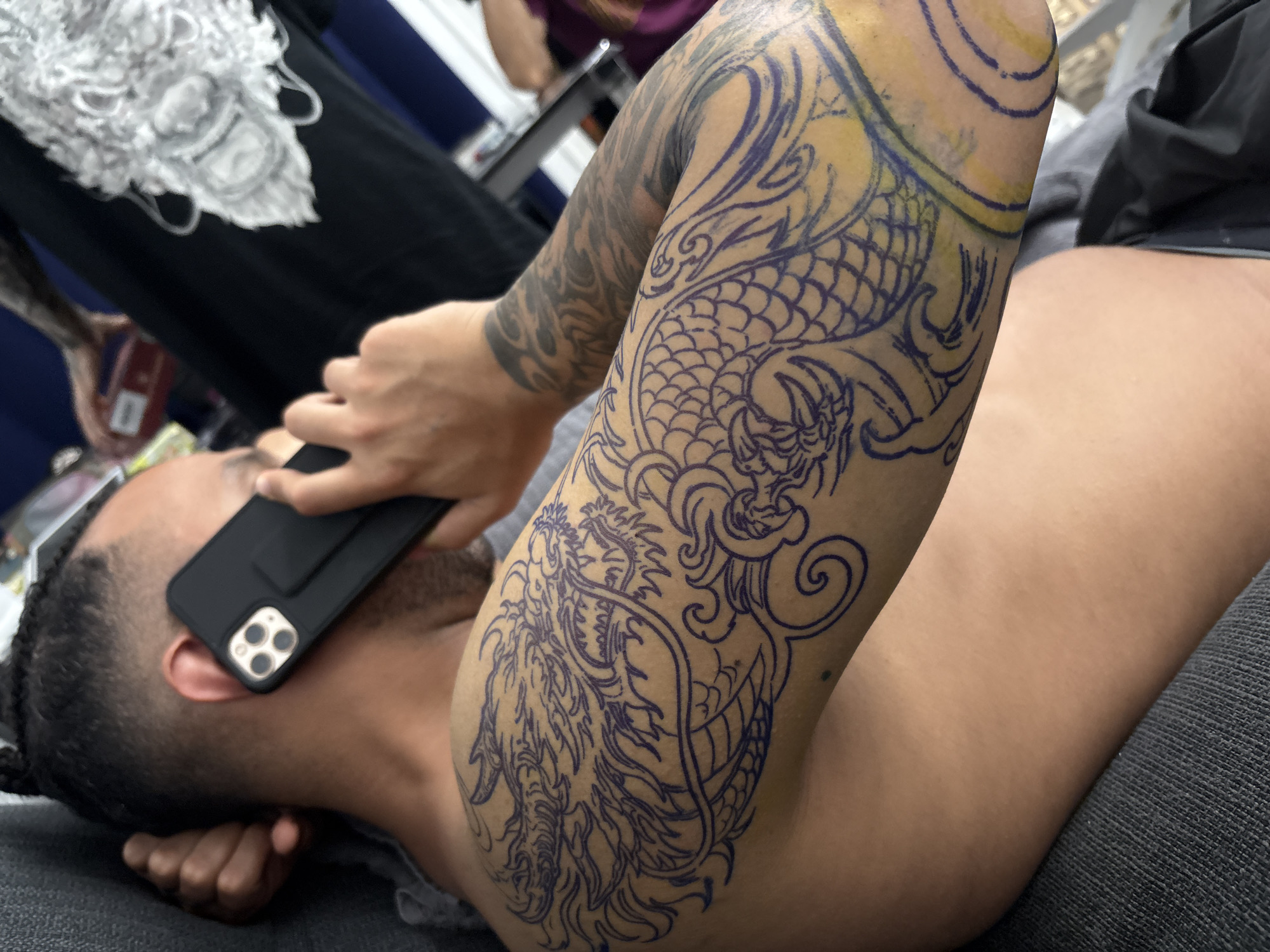 A person is lying on a grey surface, possibly a chair or a bed, while holding a black phone to their ear. The phone has a triple-lens camera and a black case with a kickstand. The person has a dragon tattoo stencil in blue ink on their upper arm. The person is wearing a grey t-shirt. In the background, there is another person wearing a maroon shirt. In the upper left, there is a white object, perhaps clothing. The scene is inside a room, likely a tattoo parlor or studio, based on the tattoo and setting. There is no evidence in this image to indicate that the scene is in Barcelona, Spain.