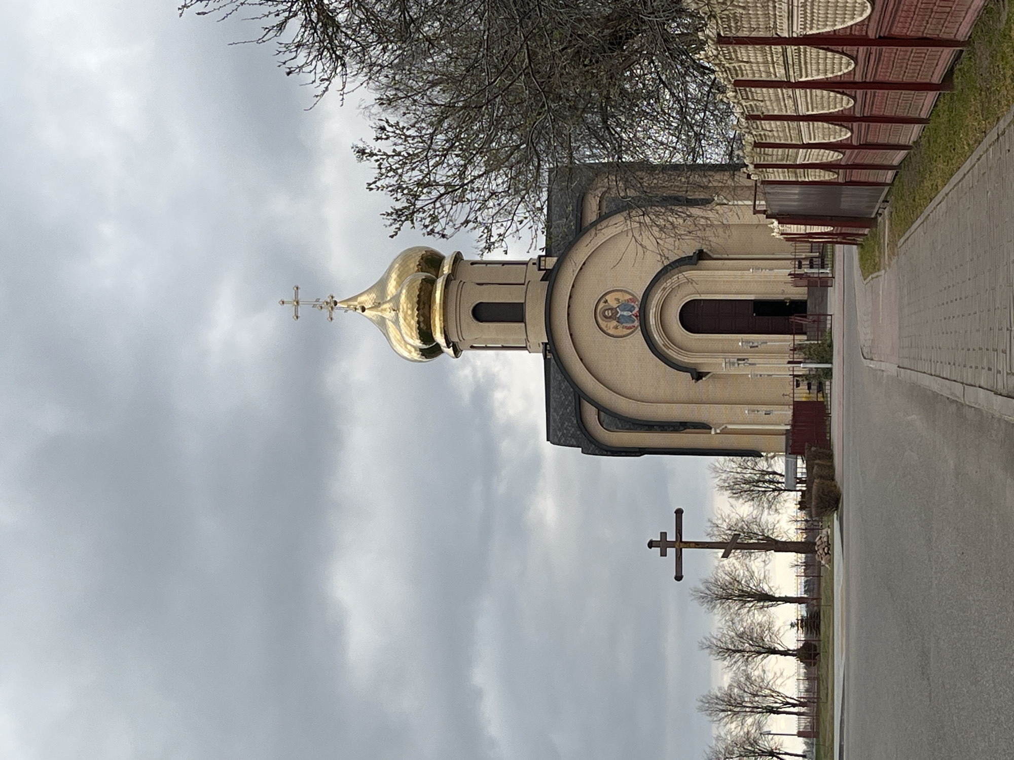 The image depicts an Eastern Orthodox church in Абухаўскі сельскі Савет, Belarus, on a cloudy day. The church features a prominent golden dome and a cross on top. The building is adjacent to a paved road. A wooden fence with a distinctive pattern runs alongside the road, parallel to the church. Further down the road, multiple large crosses stand in a row. Leafless trees are visible in the background.