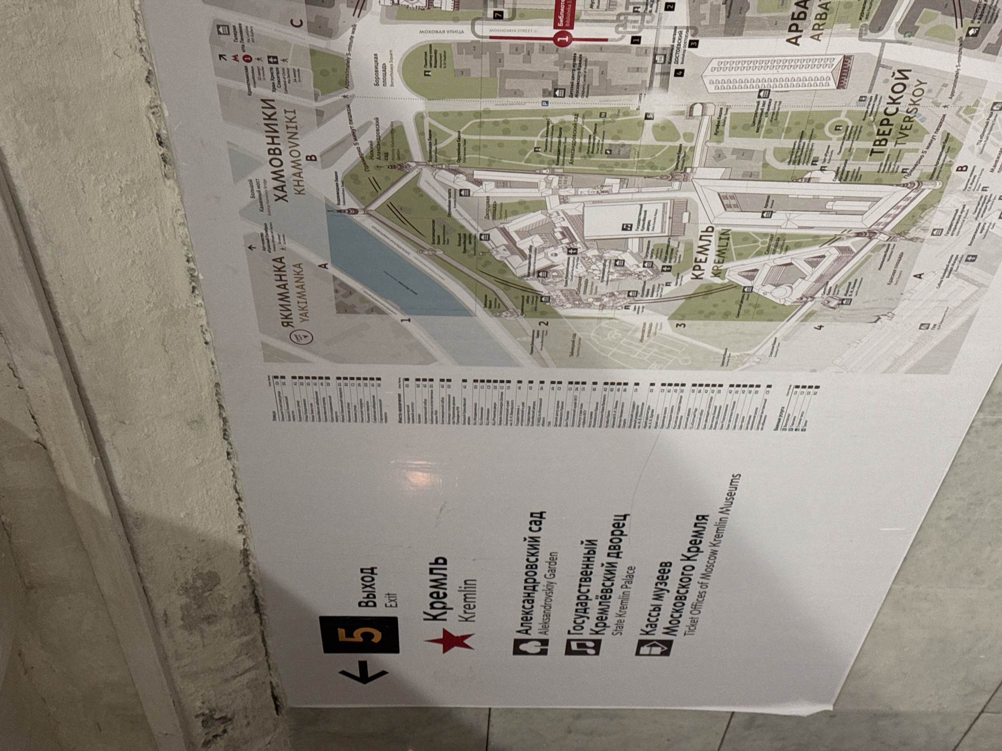 The image shows a sign with directions to various locations in Moscow, Russia. The sign is in Russian and English and points towards "Kremlin", "Alexandrovsky Garden", "State Kremlin Palace", and "Ticket Offices of Moscow Kremlin Museums". The sign also indicates an exit, numbered "5", which is to the left. A detailed map of the Kremlin and surrounding areas is displayed on the right side of the sign. The environment appears to be indoors, possibly a metro station or a public building, with a concrete ceiling visible at the top of the image. The sign is illuminated and appears to be new or well-maintained.