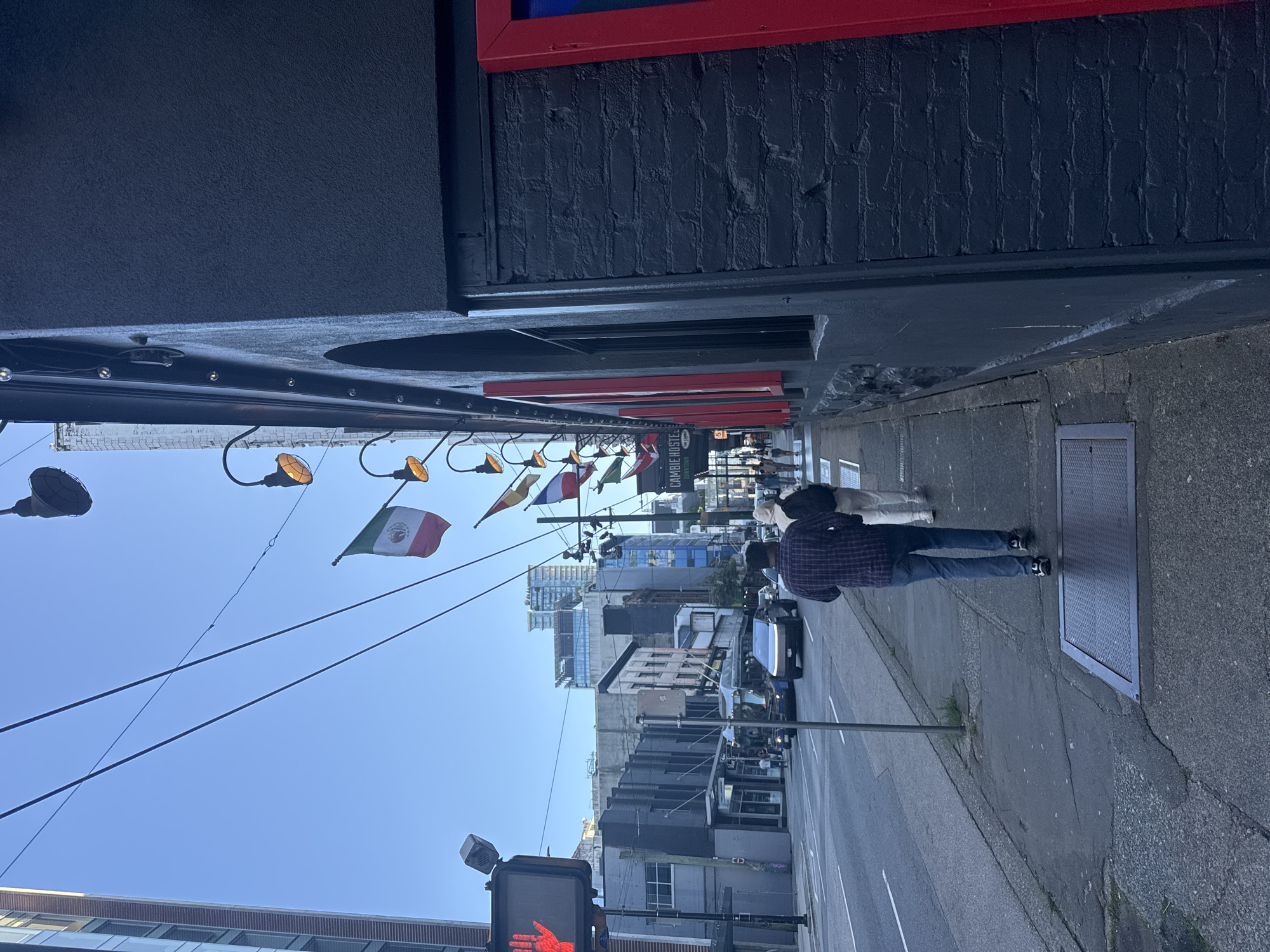 The image shows a street scene in Vancouver, Canada, likely on Cambie Street given the visible "CAMBIE HOSTEL" signage. A dark-colored, textured building with red-trimmed windows lines the right side of the frame. Flags are strung overhead, including what appears to be the Mexican flag and possibly the French flag. On the left side of the image, a sidewalk runs alongside a street with traffic, including a blue bus and a silver vehicle that resembles a Tesla Cybertruck. A pedestrian crossing signal with a red hand is visible. Several people are walking on the sidewalk, heading away from the camera. The sky is clear and blue. The overall impression is of an urban environment with a mix of modern and older architecture.