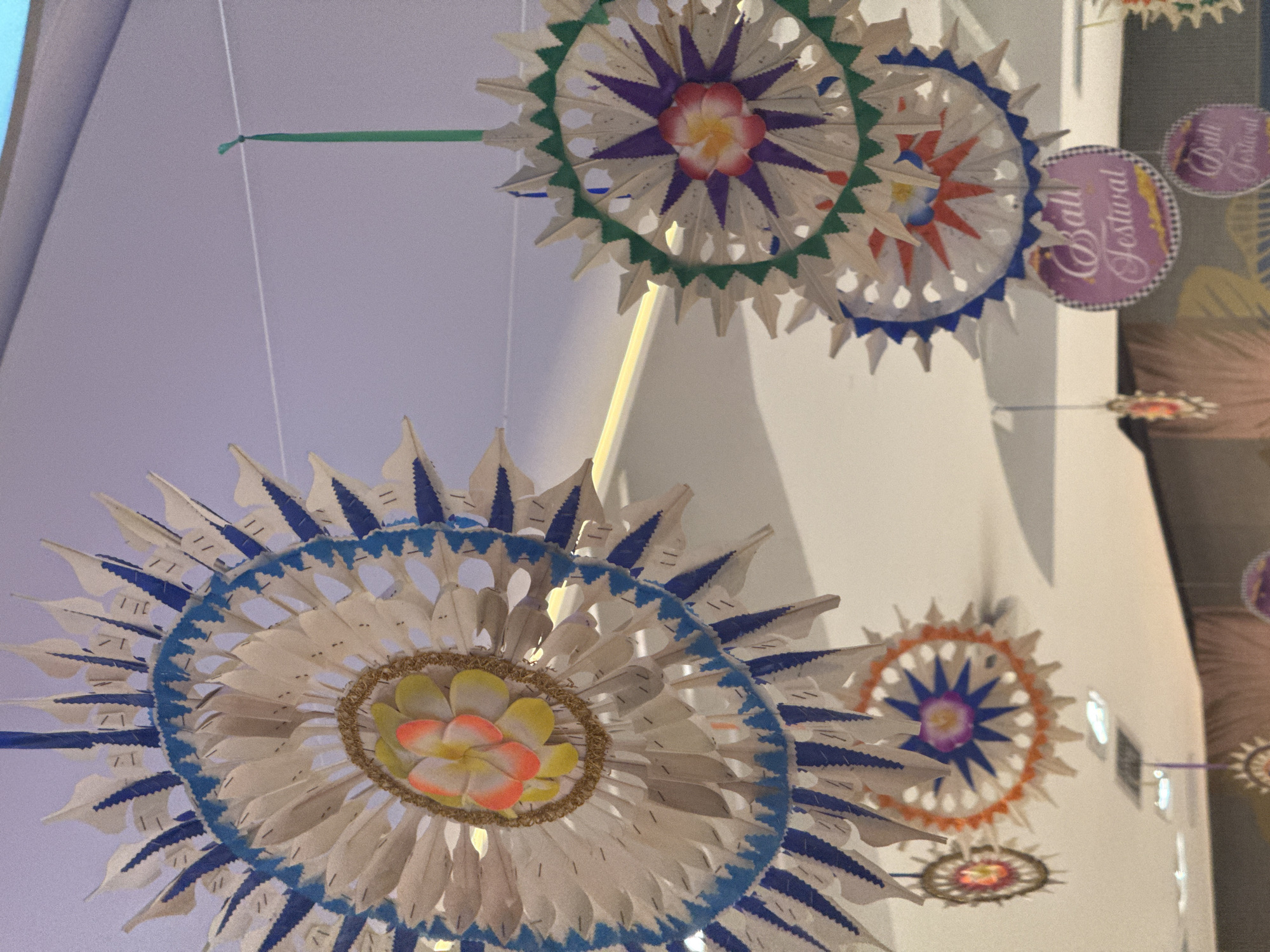 In Kuta, Indonesia, ornate, handmade decorations hang suspended from a ceiling, likely in a commercial or event space. The decorations are primarily circular, resembling starbursts or mandalas, and are crafted from white paper or thin material cut into numerous pointed segments radiating outwards. Many of these segments are edged with blue and white patterns, or solid blue. At the center of some of the larger decorations are artificial flowers, with vibrant yellow and pink petals. Smaller, simpler starbursts are visible in the background, some with orange or brown edges. Two round, pink signs with the text "Bali Festival" in a cursive font are also hanging, one below a larger decoration and another below that. The overall scene suggests a festive atmosphere, possibly related to a cultural event or market in Kuta.