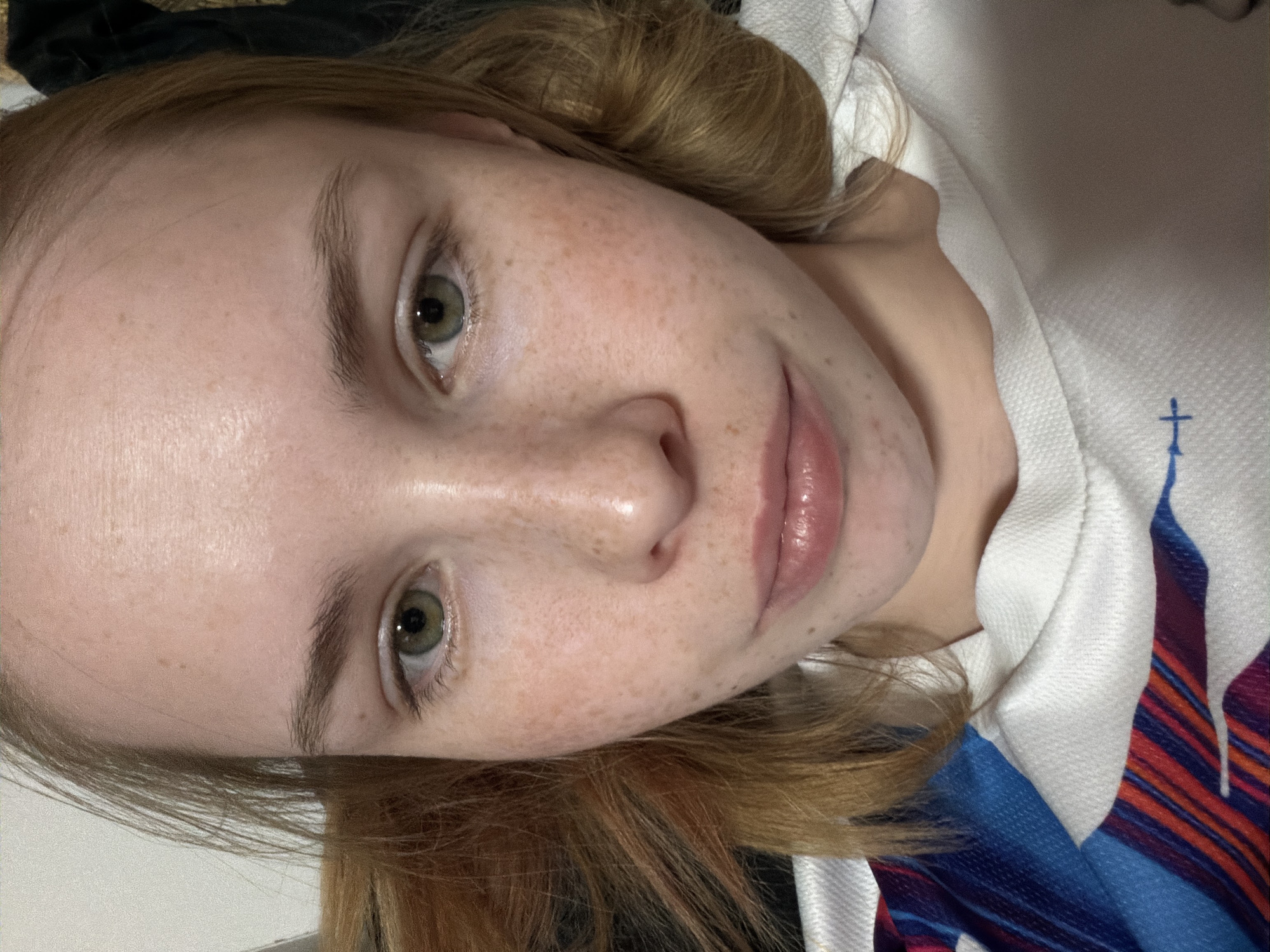 A young, light-skinned individual with freckles and green eyes is looking directly at the camera with a slight smile. Their hair is reddish-blonde and appears to be styled loosely around their head. The person is wearing a white sweatshirt that has a blue cross design on it. The background is partially visible and includes a dark area at the top left. There is no indication of the location being Berezniki, Russia.