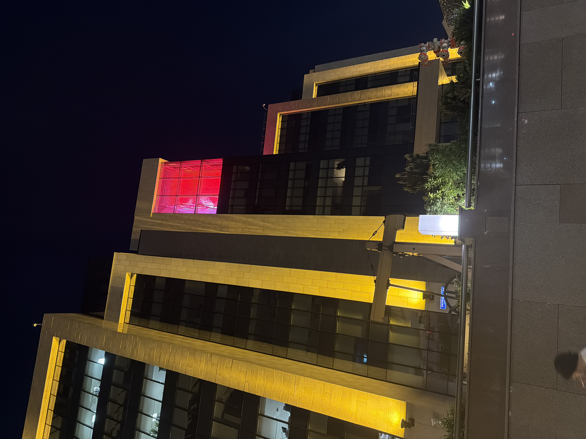 The image captures a night scene of a modern building in Moscow, Russia. The building's architectural design features a stepped or tiered structure, with each level distinctly illuminated by warm, golden light that accentuates the building's facade. The windows are dark, reflecting the night sky and the ambient light. A section of the building has a bright red illuminated feature, drawing attention to that specific area. The background is a dark night sky, contrasting sharply with the brightly lit building. The surrounding environment includes pavement, parts of other buildings, street lights, and greenery such as shrubs and trees, indicating an urban setting. A street sign and a blurry figure are visible on the pavement, suggesting pedestrian activity.