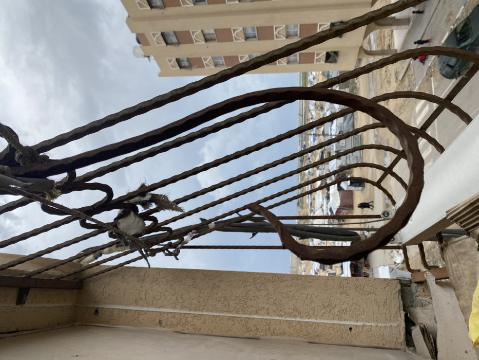 The image depicts a scene in Khan Yunis, Palestinian Territory, viewed from a slightly elevated perspective, likely a balcony or window. In the immediate foreground, a heavily rusted, dark brown wrought iron railing, featuring vertical bars and decorative curves, obstructs part of the view. Fragments of light-colored fabric or rope are entangled in the upper sections of the railing. To the far left, a section of a beige building with a textured facade is visible.
Beyond the railing, an expansive displacement camp or temporary settlement stretches across a dusty, sandy landscape. Numerous white and light-colored tents and makeshift shelters are densely arranged. A narrow dirt path or road traverses the camp. On this path in the mid-left, two individuals walk together, one wearing a dark garment. Further to the right and closer to the foreground, a small group of approximately three individuals, including at least one child in a red top, are visible on a street. A dark-colored SUV is partially visible on this street in the lower right.
In the background, on the far right, a multi-story, light brown building with multiple rectangular windows and distinct arched decorative elements above the windows on each floor stands prominently. A white water tank is visible on its roof. Beyond the camp, a distant dark line suggests a perimeter wall or boundary. The sky above is overcast with a mix of light and dark grey clouds.