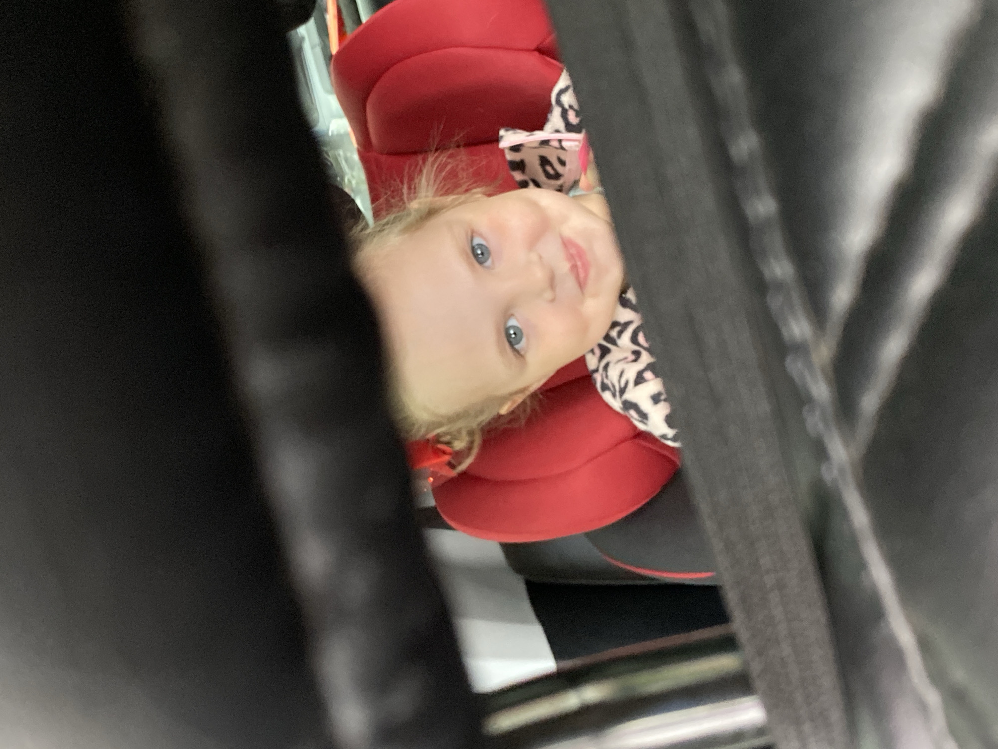 A close-up shot focuses on a young girl with blonde hair and blue eyes, seated in a red and black car seat. She is wearing an animal print top. The image is framed by dark, blurred elements on either side, obscuring the full view of the surroundings. Due to the obscured perspective and lack of visible landmarks, it's not possible to definitively confirm the location as Votkinsk, Russia, or any other specific place.