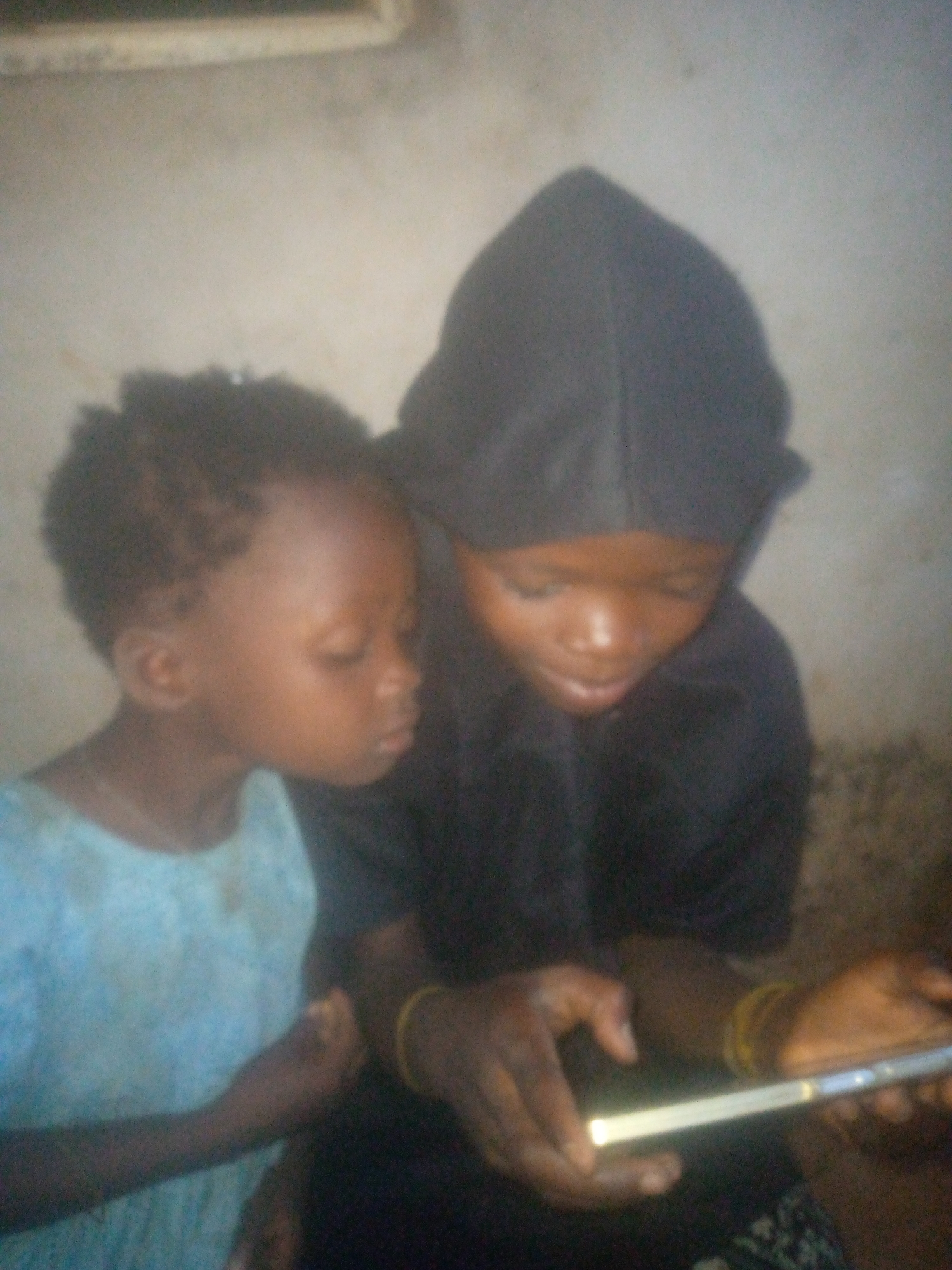 Two children are closely watching a brightly lit cellphone screen. The child on the left is wearing a light blue shirt. The child on the right is wearing a dark hooded shirt and has gold bracelets on their wrist. The location is not discernable.