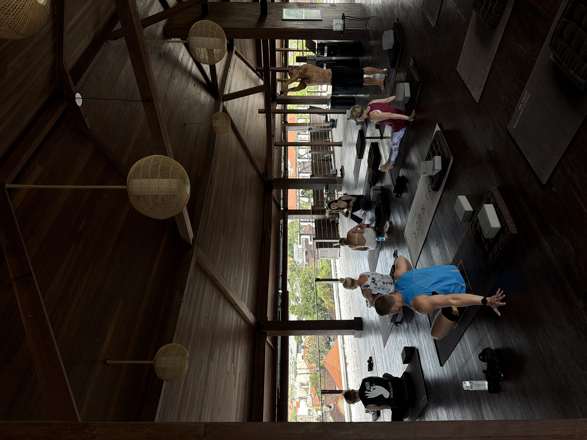 This image captures an indoor yoga class taking place in a spacious, open-air studio with a wooden ceiling and floor, overlooking the rooftops of Mengwi, Indonesia. The room is furnished with yoga mats, blocks, and bolsters, and illuminated by natural light filtering in and hanging woven pendant lights. Several individuals are engaged in yoga poses on their mats. In the foreground, a man in a blue tank top is in a seated yoga pose, with his left hand on the floor for support. To his right, a woman in a maroon tank top and patterned leggings is also seated on her mat, reaching forward. Towards the center of the room, a man with tattoos on his torso, wearing black shorts, stands and appears to be instructing the class. Other participants are visible in various seated positions around the studio. The background reveals a panoramic view of the urban landscape of Mengwi, with terracotta-tiled roofs and lush green foliage.