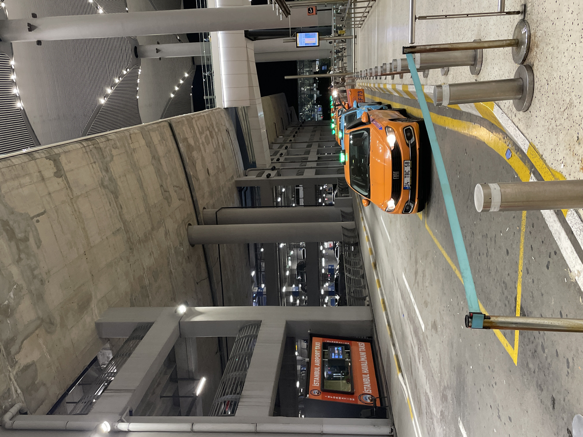 The image depicts a taxi stand at Istanbul Airport, located in Arnavutköy, Turkey. The scene is set outdoors, beneath a vast concrete overpass structure and the futuristic architecture of the airport terminal building. A line of taxis, predominantly orange and blue, are queued on the asphalt roadway, their taxi lights illuminated green. The foreground features yellow and white road markings and a bright turquoise ribbon barrier. To the left, an orange kiosk labeled "ISTANBUL AIRPORT TAXI" and "ISTANBUL HAVALIMANI TAKSI" is visible, providing information. The background shows multiple levels of a parking garage with cars visible and artificial lighting. The overall atmosphere is that of a busy transportation hub.