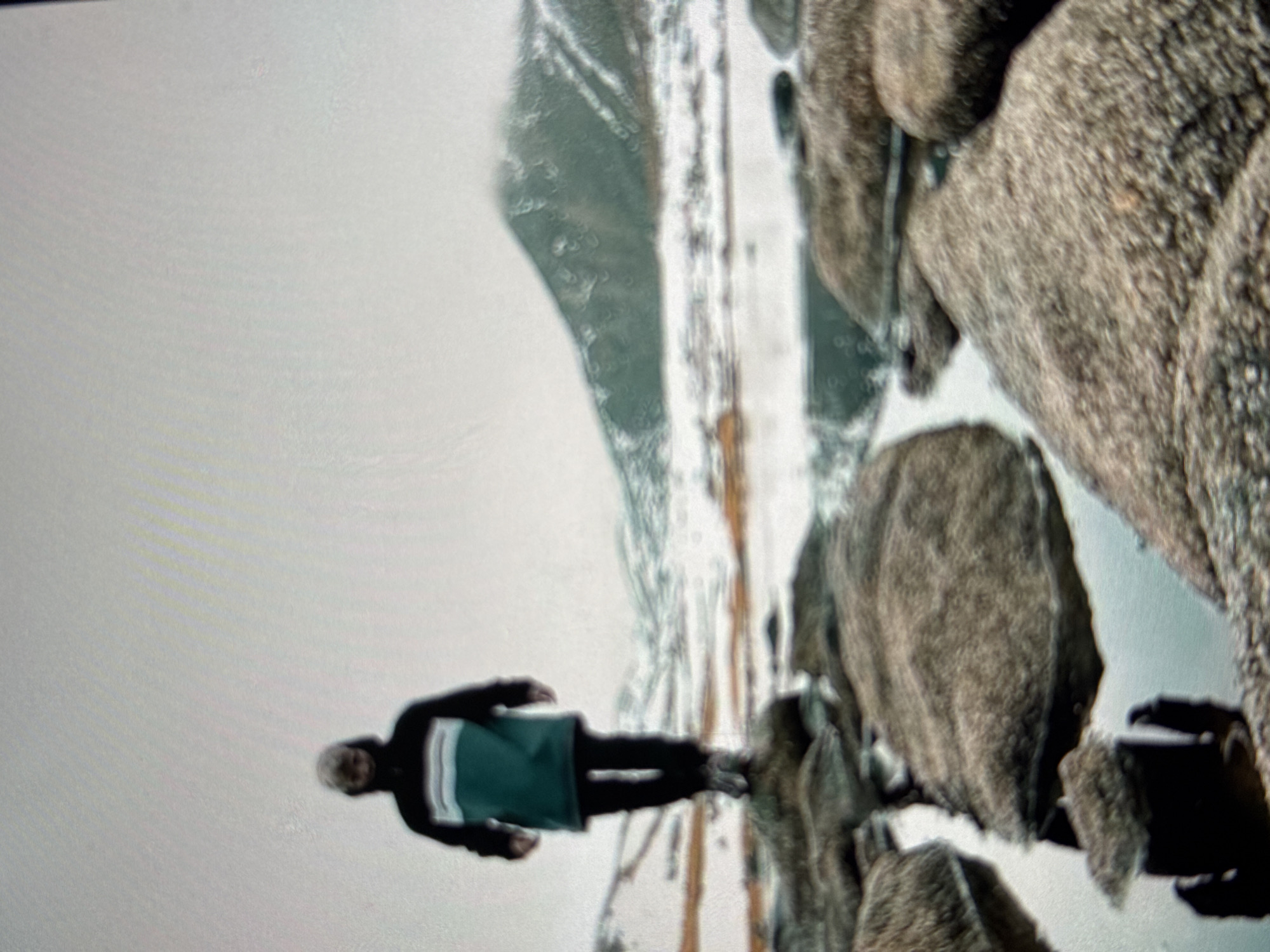 A full shot captures a person standing on a rock in a body of water. The person is facing away from the camera, wearing a dark jacket with green and white stripes on the chest, black pants, and gray boots. The environment is mountainous and appears to be covered in snow. The water in the foreground reflects the person's shadow. The sky is overcast. The image's resolution is low, and there are water spots on the surface. This scene is not located in Irkutsk, Russia.