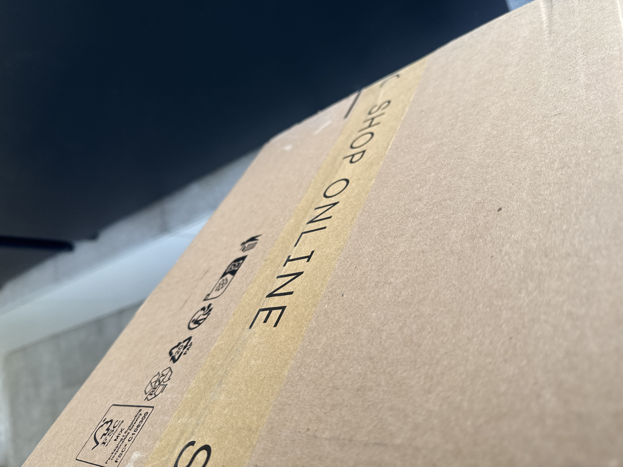 The image shows a close-up of a cardboard box. The box is printed with the words "SHOP ONLINE" in black block letters, running vertically along the side. The words appear to be printed on a tan-colored tape that is affixed to the box. On the left side of the tape, there are several small recycling symbols and an FSC certification logo. The location is unknown.