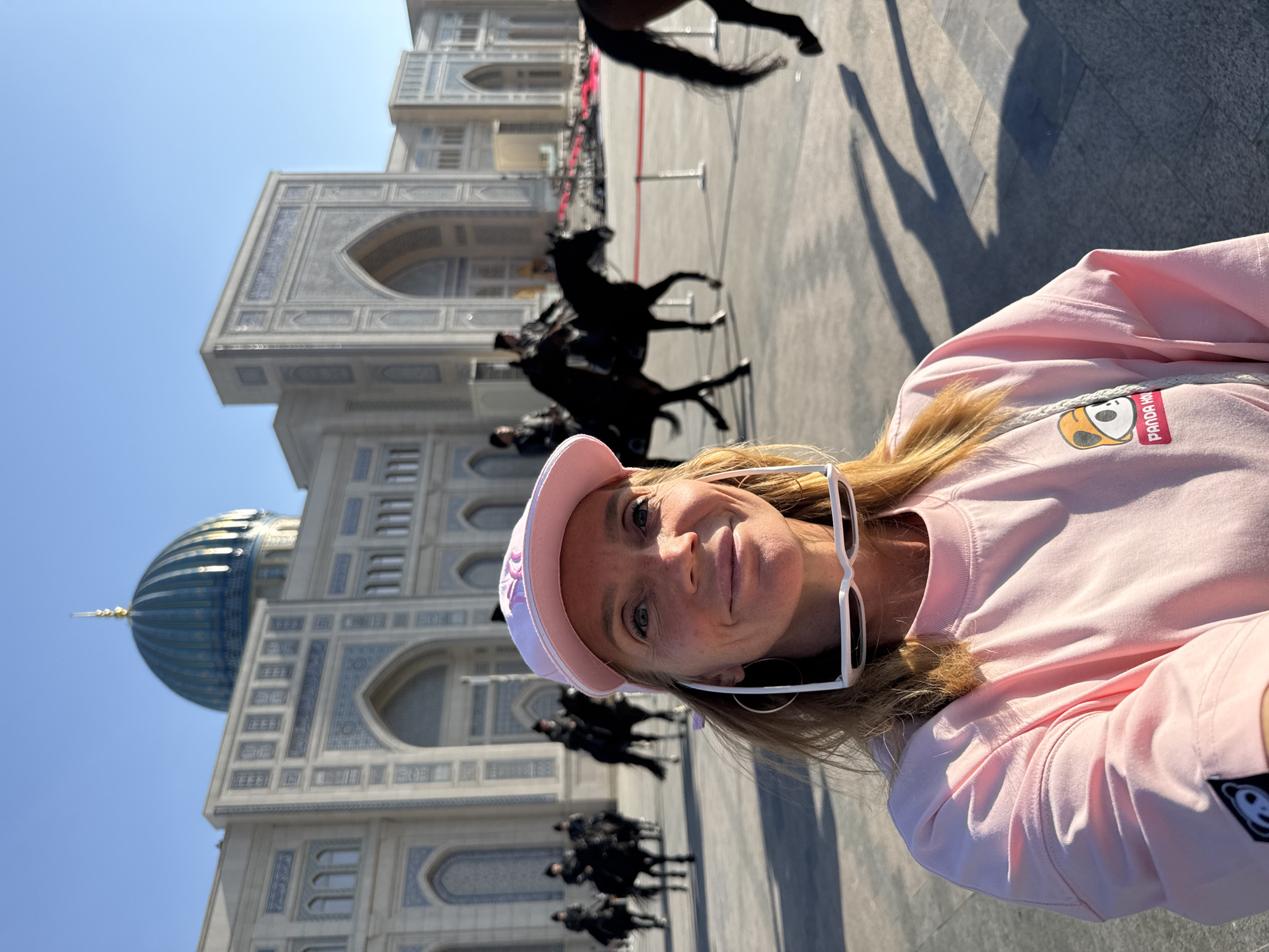 A woman in a pink hat and sweatshirt takes a selfie in front of the Palace of International Forums in Tashkent, Uzbekistan. The Palace is a large, ornate building with a blue dome and intricate architectural details. In the background, a line of riders on horseback is visible. The sky is clear and blue. The woman is wearing white sunglasses and hoop earrings. Her sweatshirt features a panda bear design. The image is rotated.